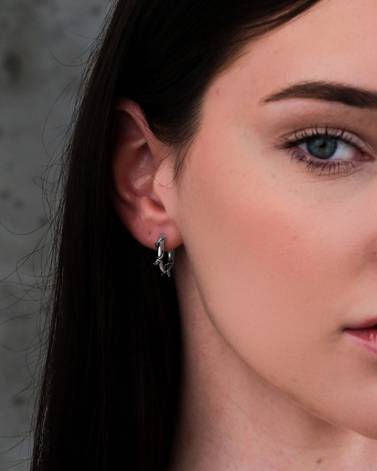 Close-up of a woman with fair skin, long dark hair, and blue eyes in profile, showcasing her ear adorned with Barbed Wire Hoop Earrings in stainless steel. She has a neutral expression against a grey background.