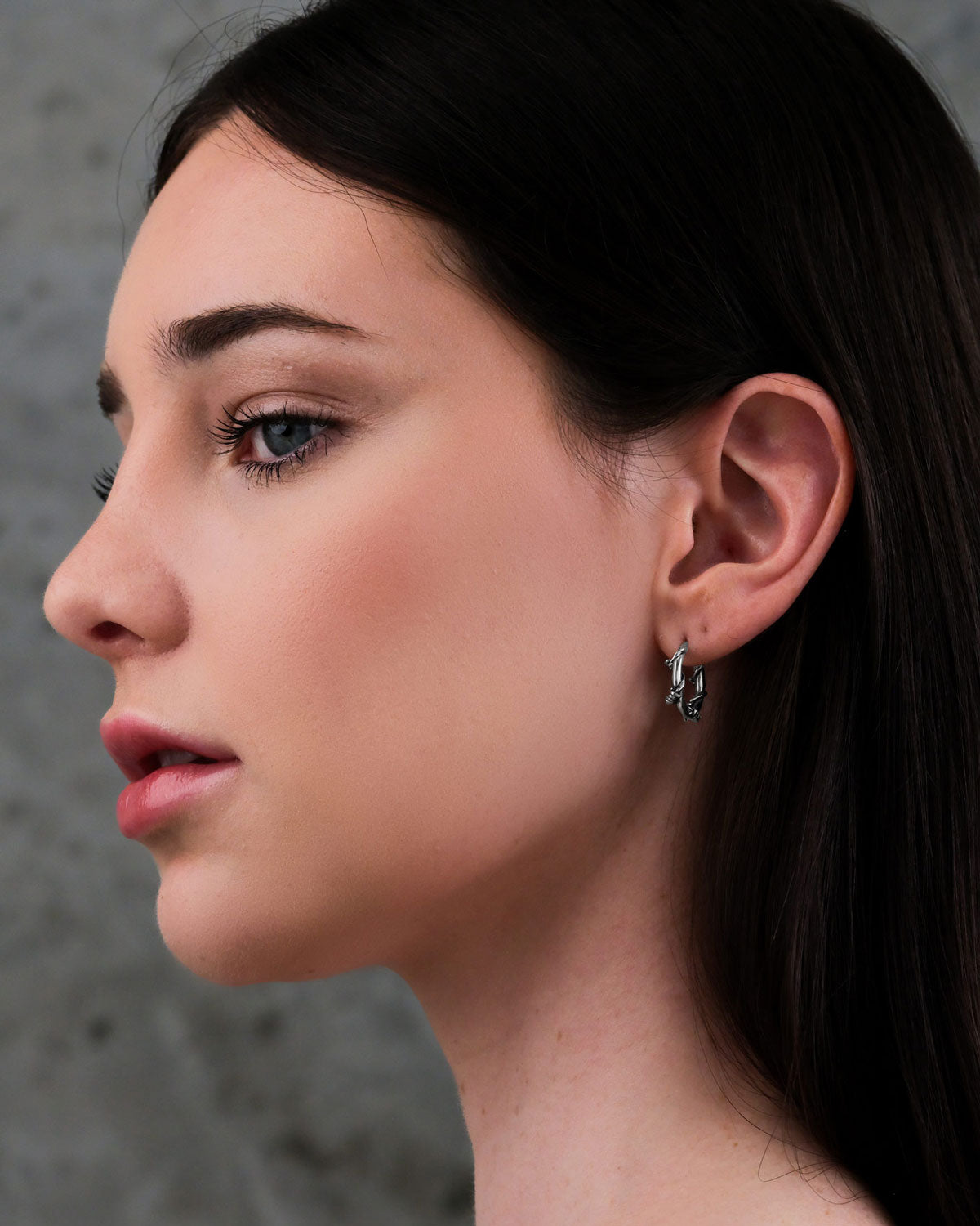 A woman with long dark hair and fair skin is shown in profile against a gray background, wearing subtle makeup and Barbed Wire Hoop Earrings.