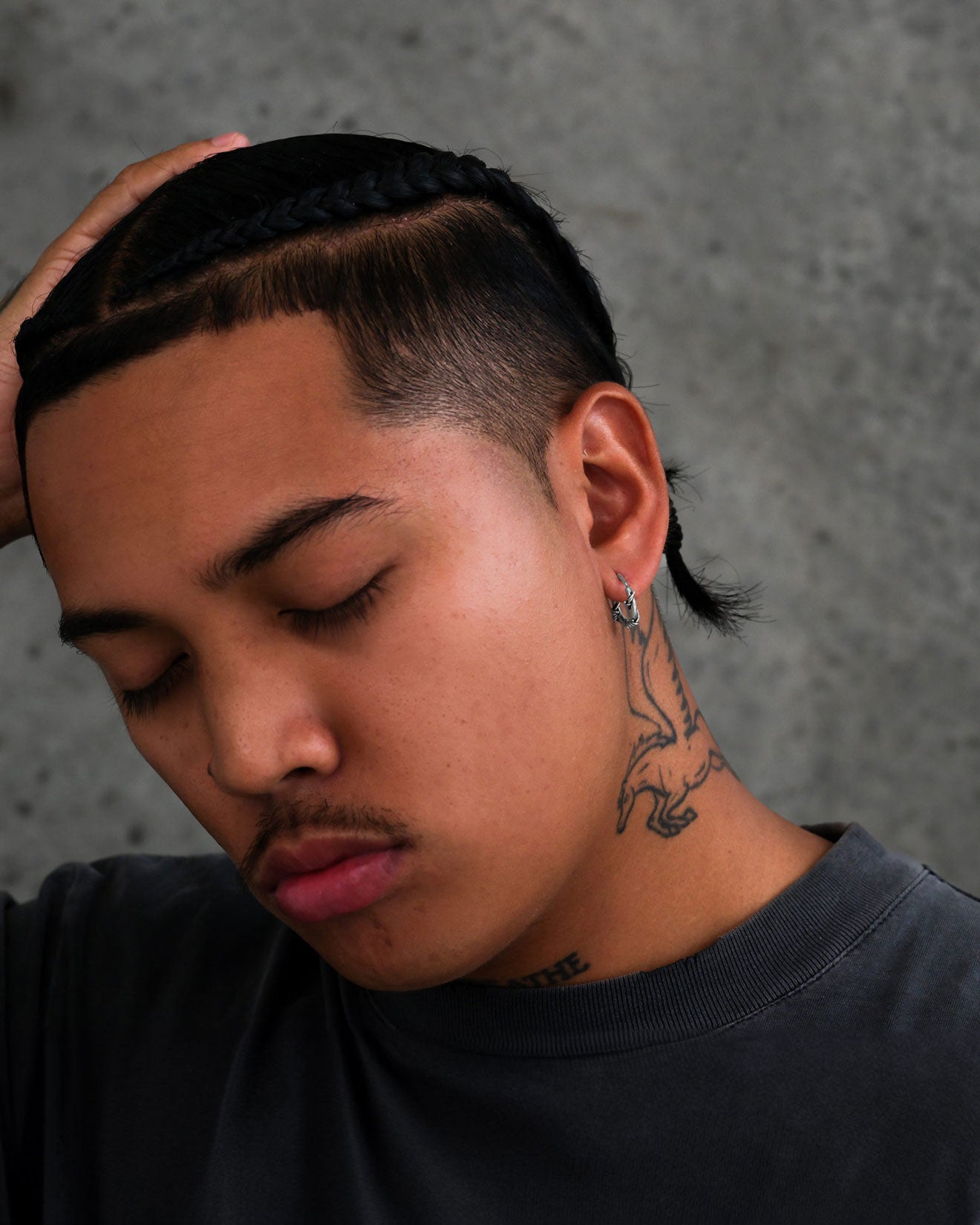 A person with braided hair, closed eyes, and a faint mustache touches their head. They wear a dark shirt and Barbed Wire Hoop Earrings, showing off a dinosaur neck tattoo against a textured gray background.