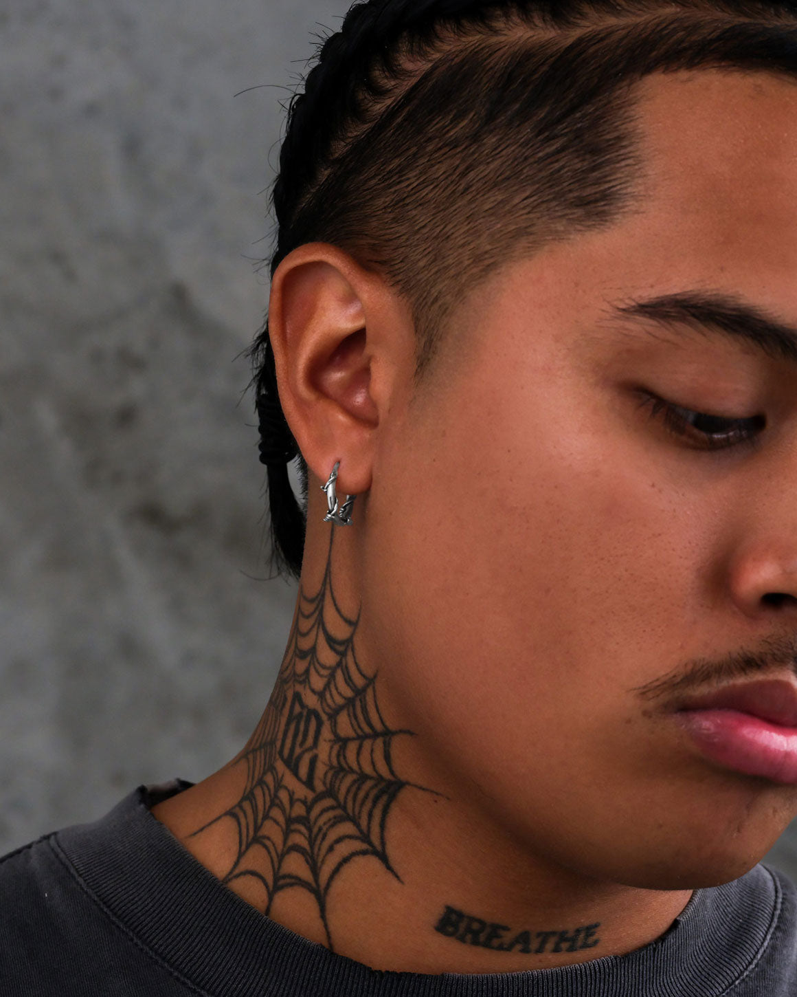 A person with braided hair and a mustache wears small Barbed Wire Hoop Earrings. A spiderweb tattoo decorates the neck, with "BREATHE" inked below. The background is a gray, textured wall.