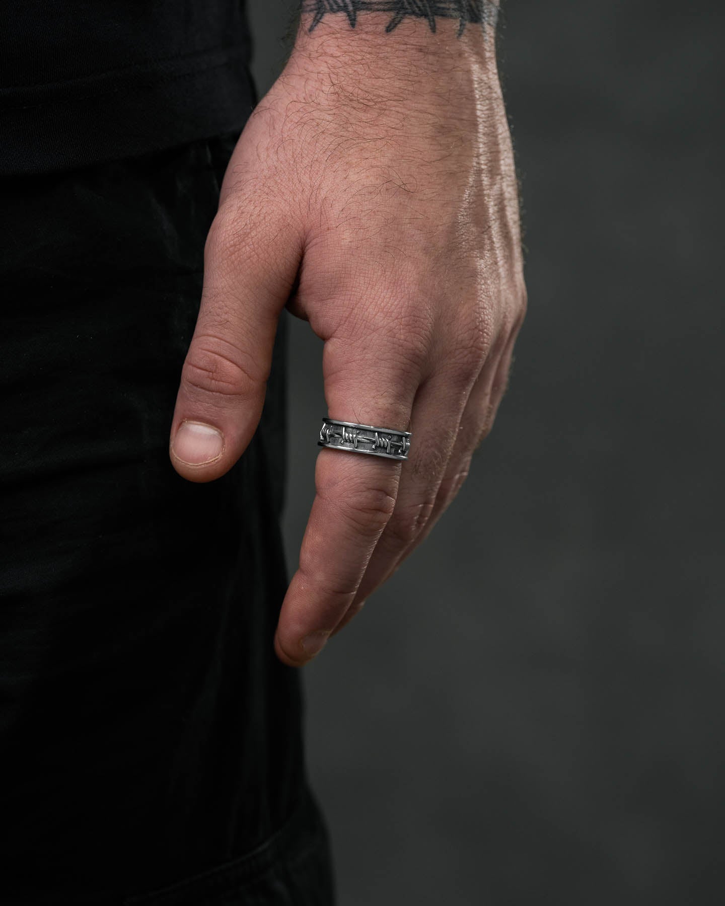 A close-up of a hand wearing the Barbed Wire Ring, featuring engraved designs. The hand has visible veins and a wrist tattoo, set against a dark, blurred background.