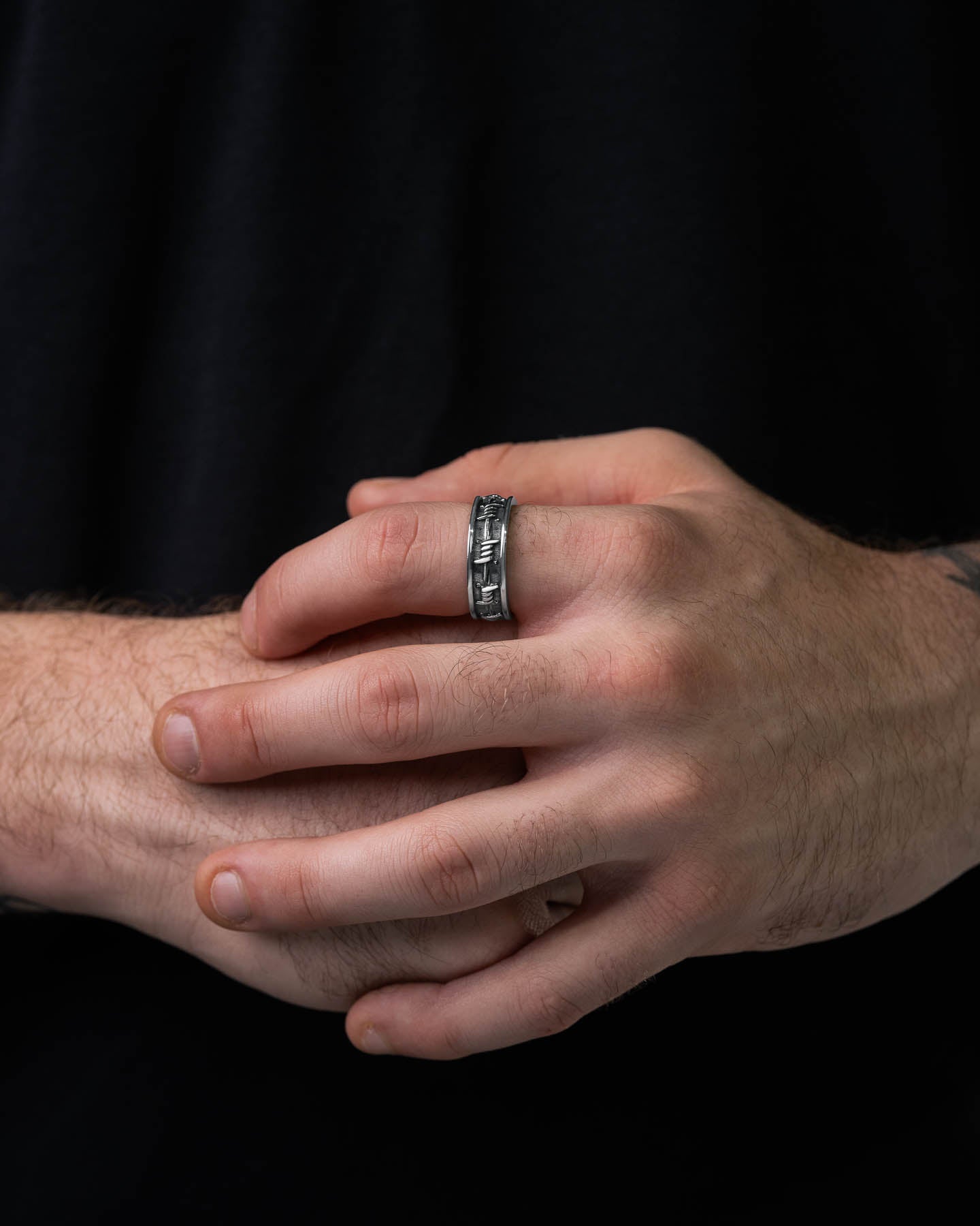 A close-up of hands, one gently holding the other, wearing the Barbed Wire Ring—a stainless steel band with a textured design—on the ring finger. The person is in a dark shirt against a black background.