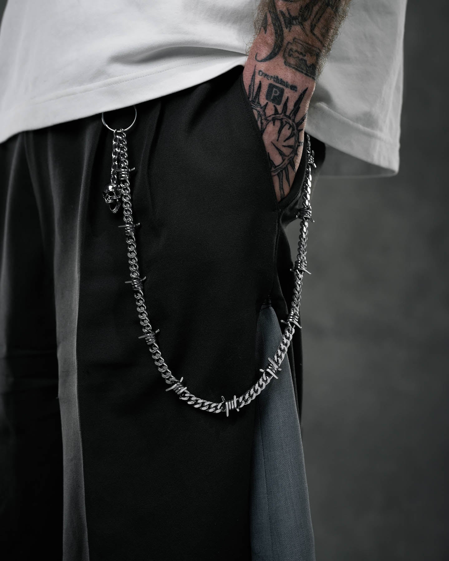 A person wearing black pants with a 9mm Barbed Wire Cuban Wallet Chain hanging from the pocket. Their tattooed arm is by their side, set against a dark, blurred background that creates a moody vibe.