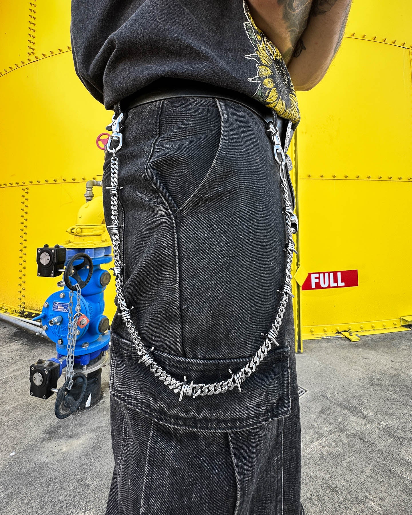 A person wearing black jeans with a 9mm Barbed Wire Cuban Wallet Chain stands near a bright yellow industrial tank marked FULL, with blue valves and pipes in the background.