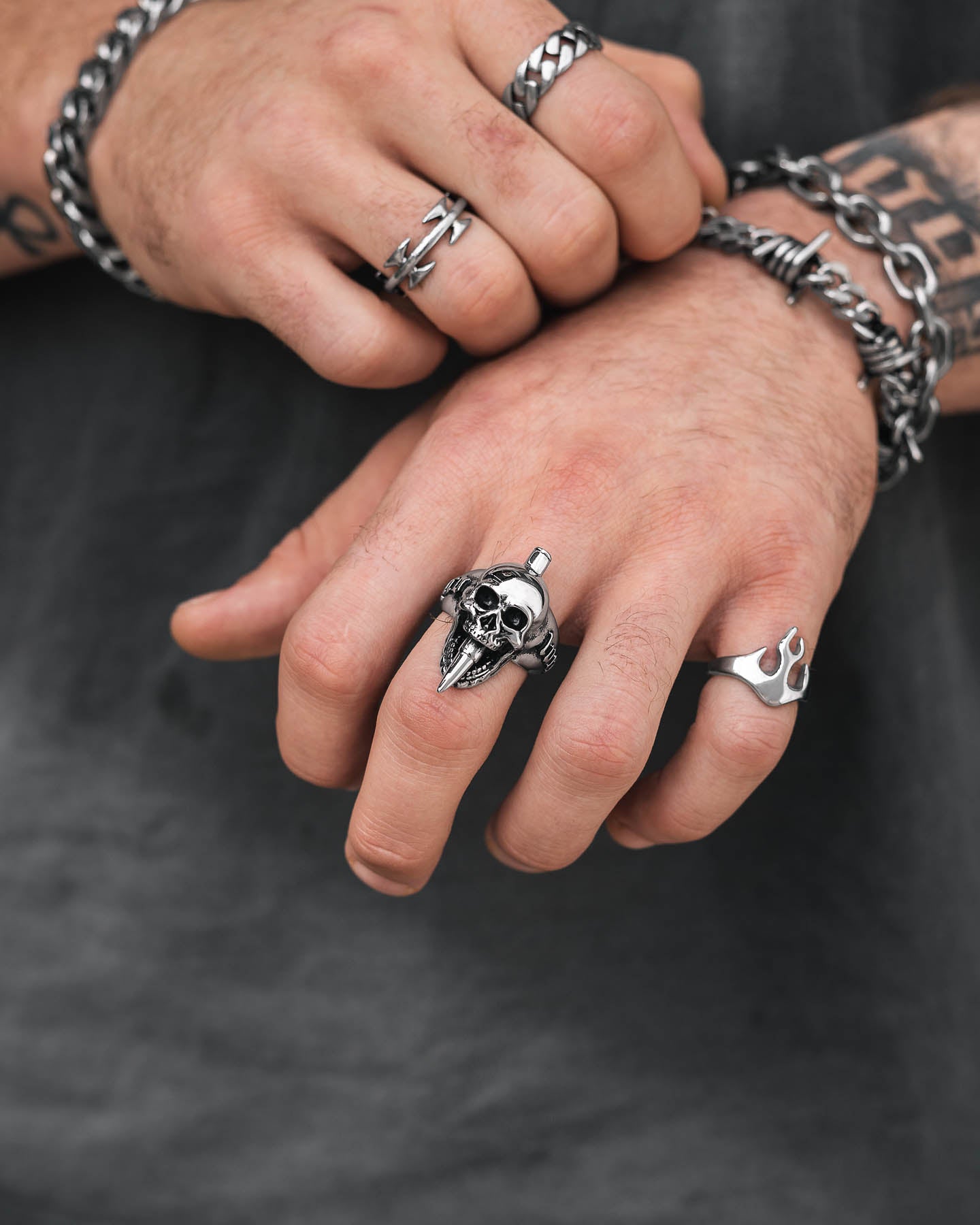 Close-up of two hands with tattoos and wearing multiple silver rings and bracelets, including the "Bite The Bullet" Ring. The person is also dressed in a dark shirt, highlighting their edgy rock-inspired style.