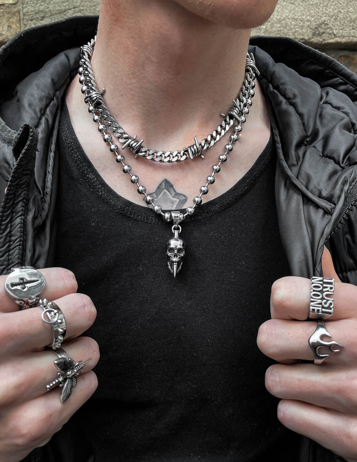 A person wears layered silver chains, including the "Bite The Bullet" Pendant and a skull necklace, plus silver rings with unique designs. They sport a black tank top, dark jacket, and display a geometric chest tattoo.