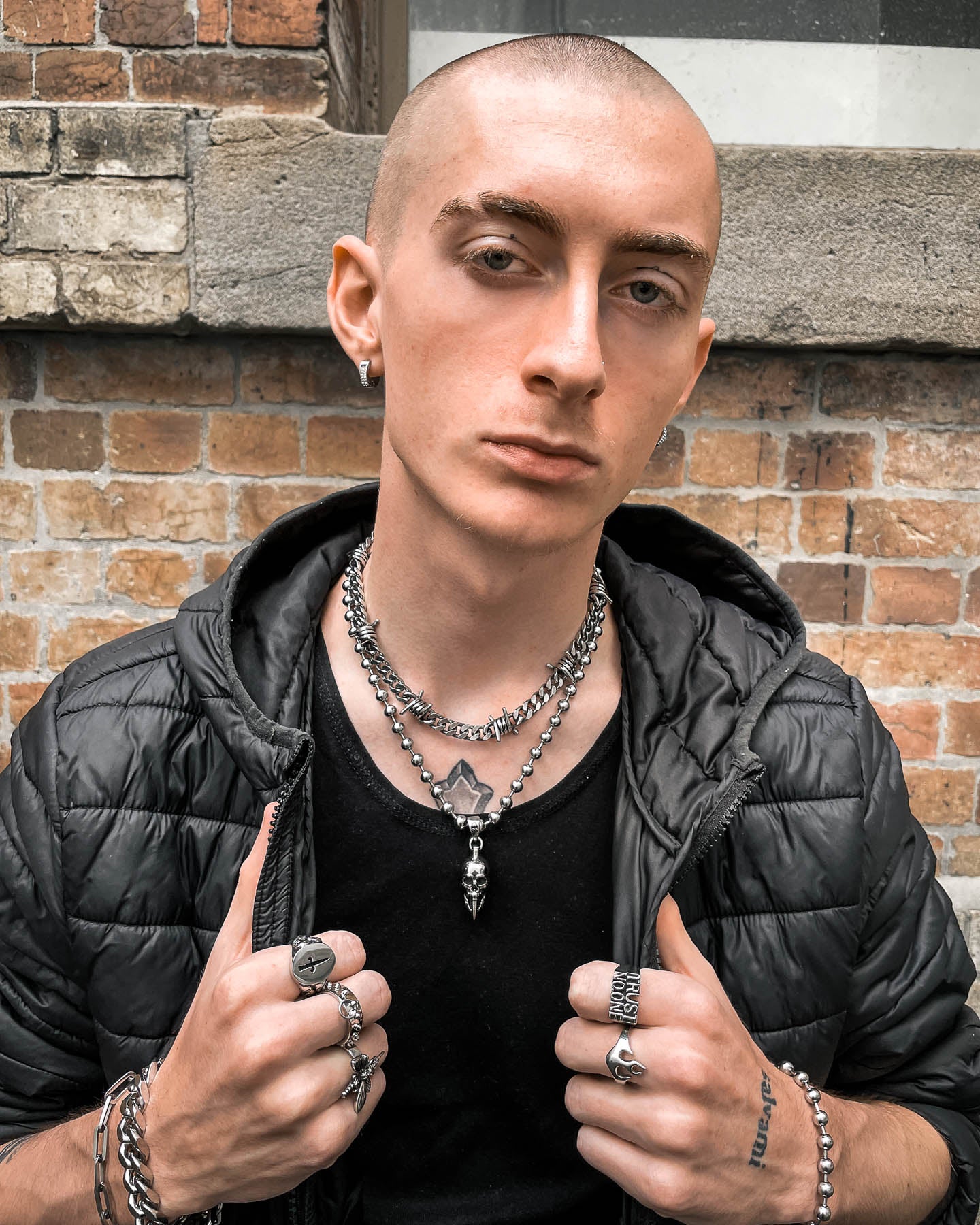 A person with a shaved head and eyebrow piercing wears layered silver necklaces featuring the "Bite The Bullet" Pendant, along with rings, hoop earrings, a black shirt, and jacket, standing confidently before a brick wall.