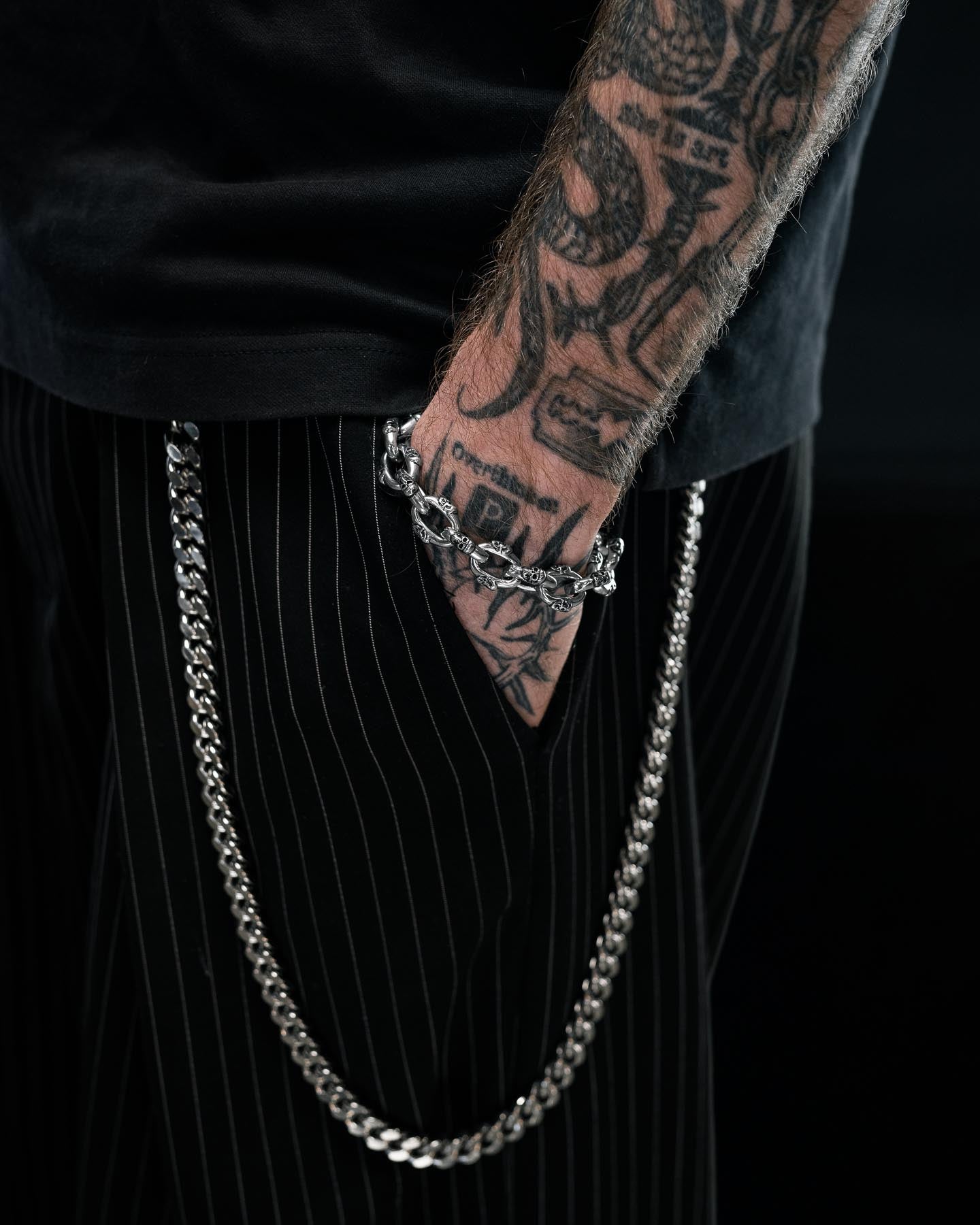 A tattooed arm rests by black pinstripe pants with a silver chain hanging from the waistband, wearing the Statement Collective "Boneyards" Skull Bracelet on the wrist, set against a dark background.