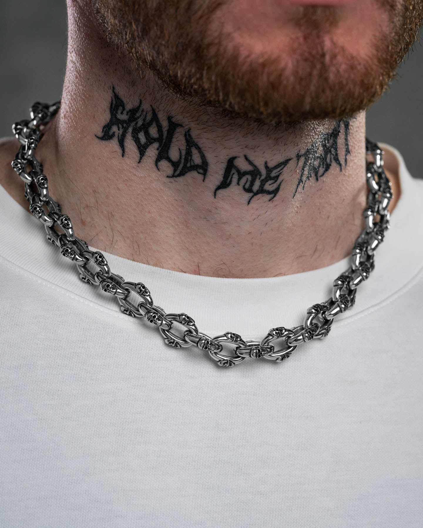 A close-up of a man's neck with a black Could Not Wait tattoo in stylized script and the "Boneyards" Skull Chain, a chunky stainless steel necklace. He wears a white shirt, and part of his beard is visible.