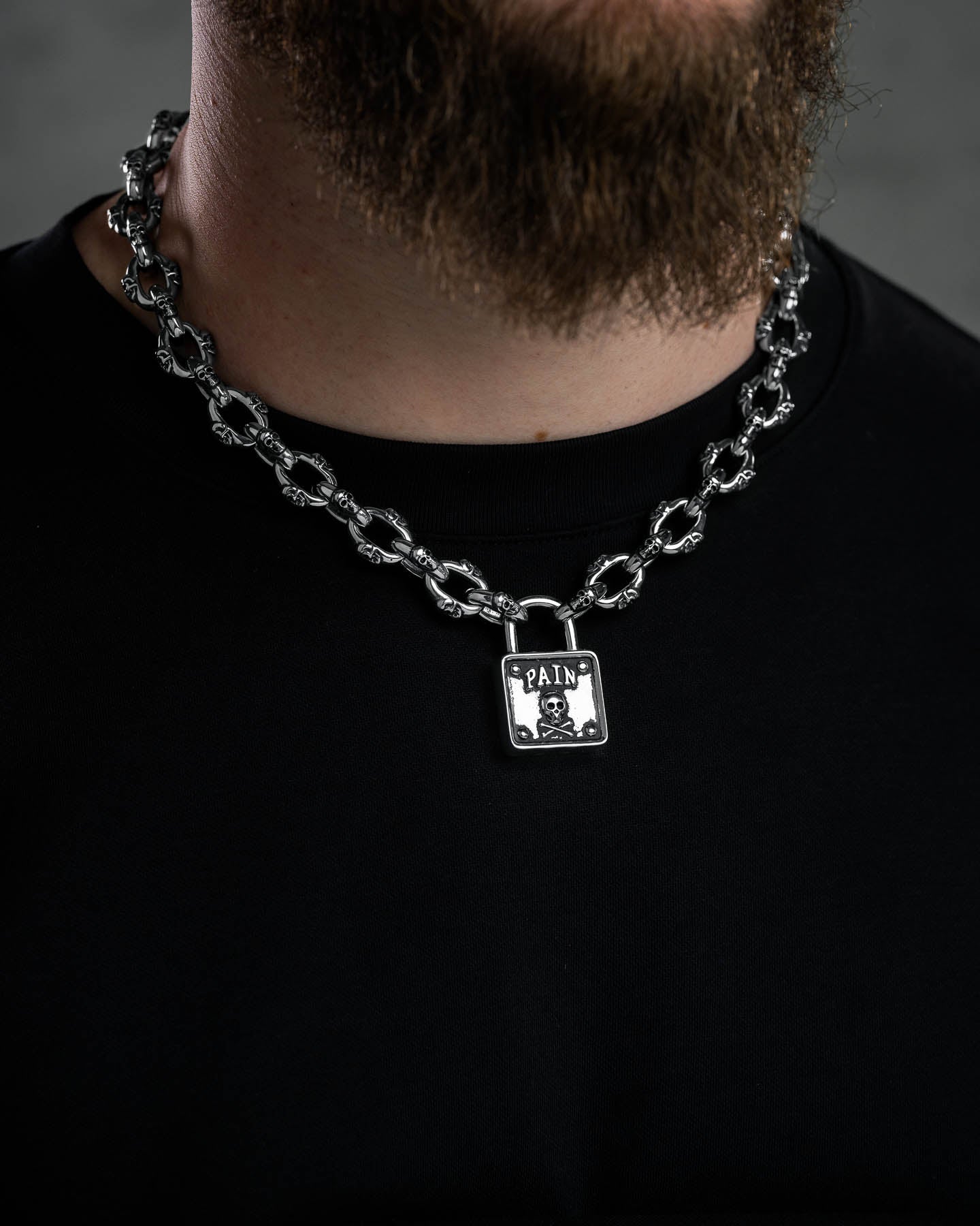 A person wears the "Boneyards" Skull Padlock Chain—12mm stainless steel cable chain featuring a square pendant with "PAIN" engraving and a small skull—paired with a plain black shirt.