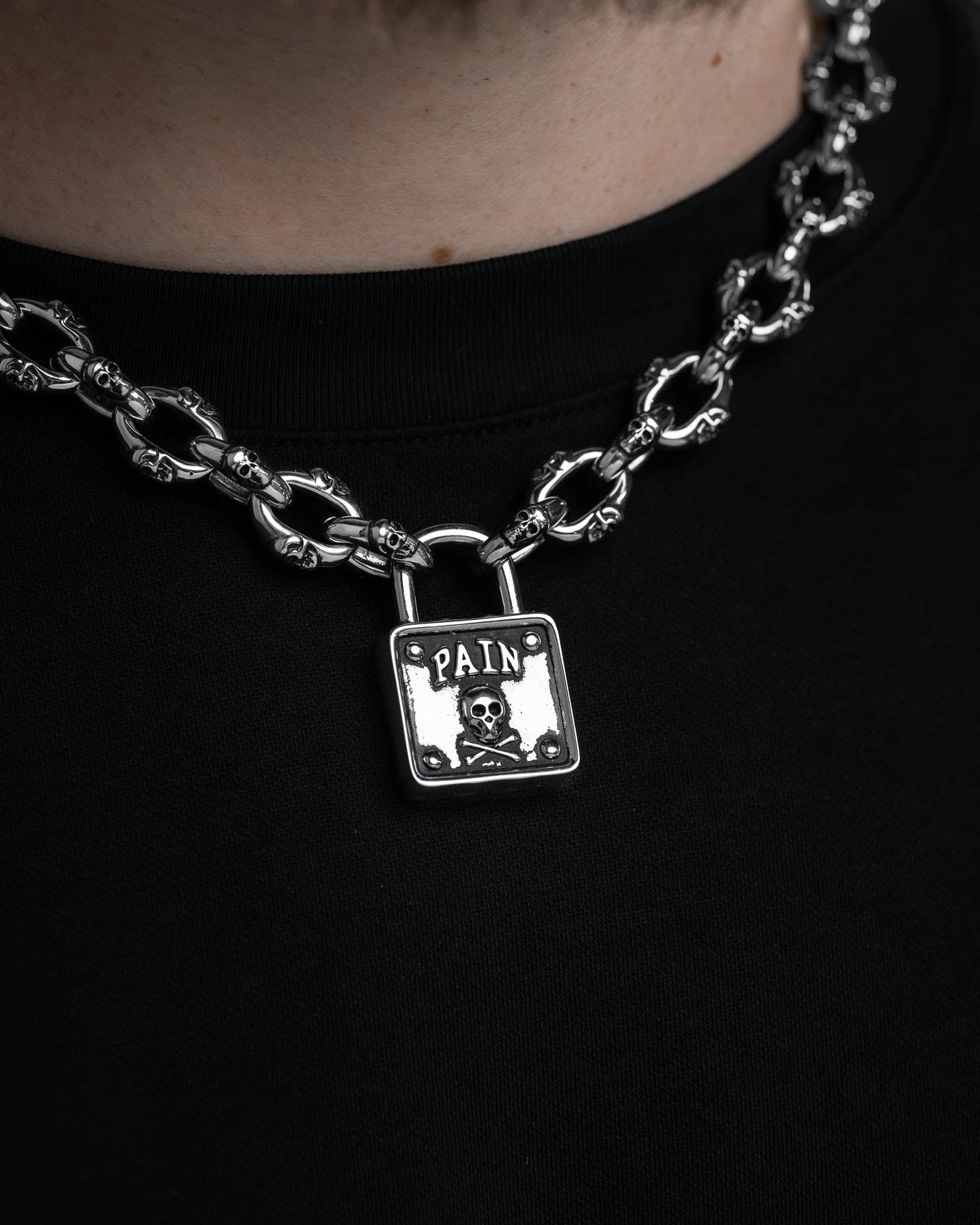 A close-up of the "Boneyards" Skull Padlock Chain—a stainless steel necklace with 12mm cable chain, silver skull links, and a large PAIN-engraved padlock pendant with a skull and crossbones, styled over a black shirt.