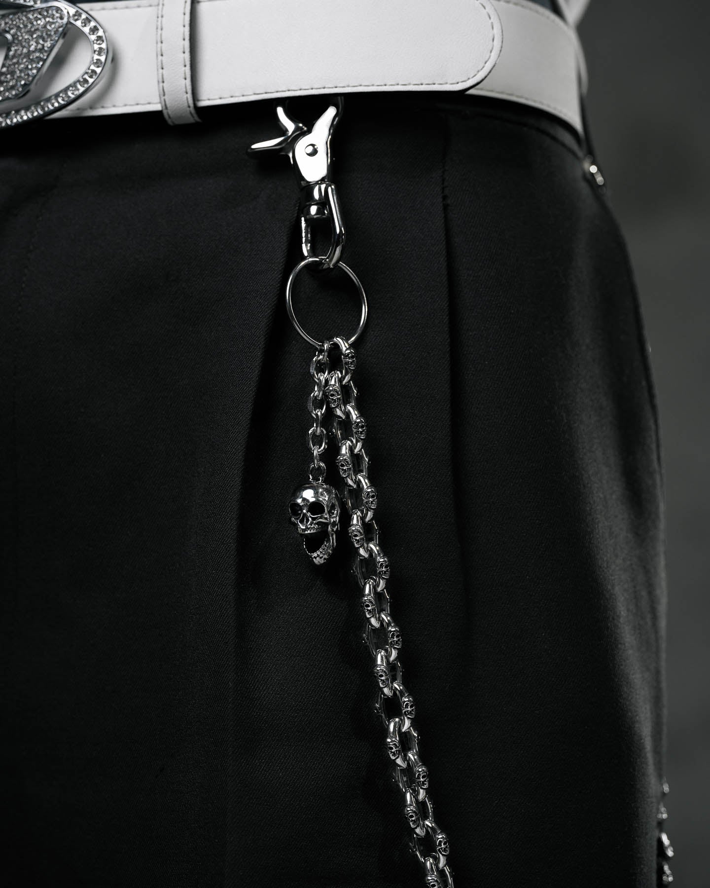 Close-up of black pants with the "Boneyards" Skull Wallet Chain, showcasing a silver skull charm attached to the waistband by a white belt with a shiny decorative buckle and clasp.