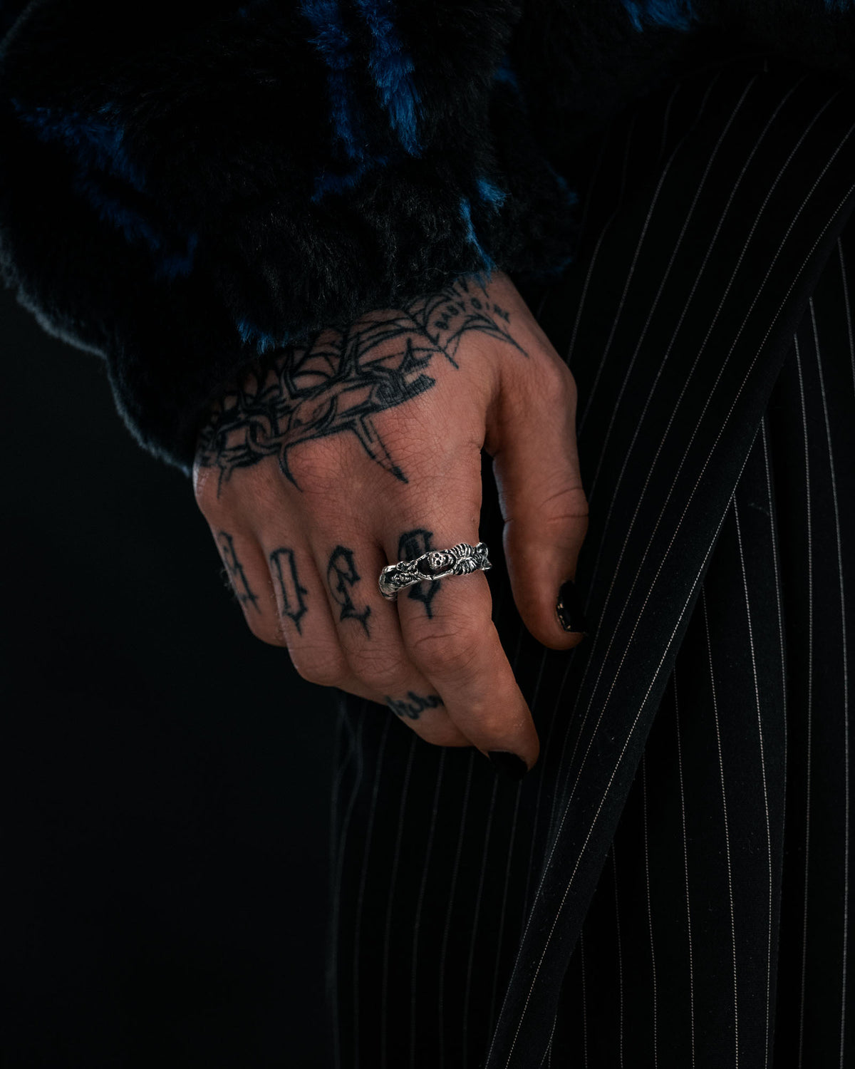 A tattooed hand with black-painted nails wears the Breaking Wheel Ring by Statement Collective and rests against black pinstriped pants. The person is wearing a fuzzy dark sweater with blue accents.