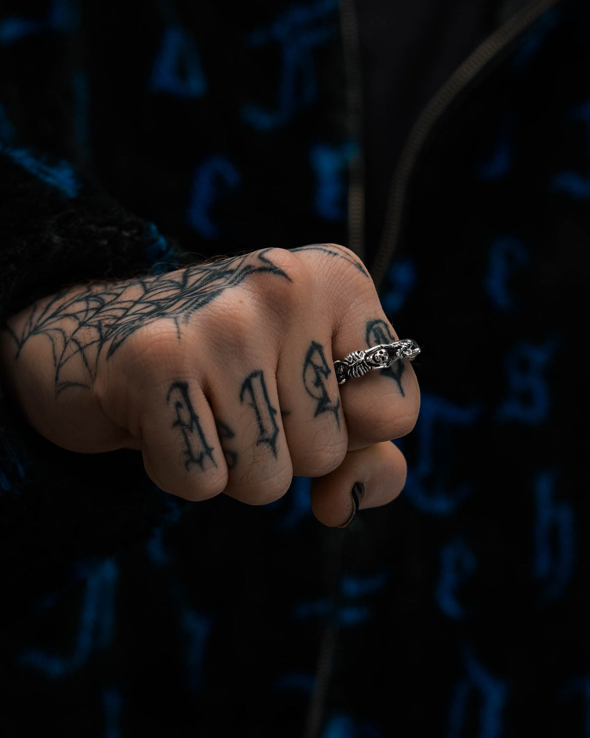 A tattooed hand, featuring a spider web and letters on the fingers, wears the Breaking Wheel Ring by Statement Collective—a detailed silver piece—paired with a dark jacket adorned with a blue pattern.