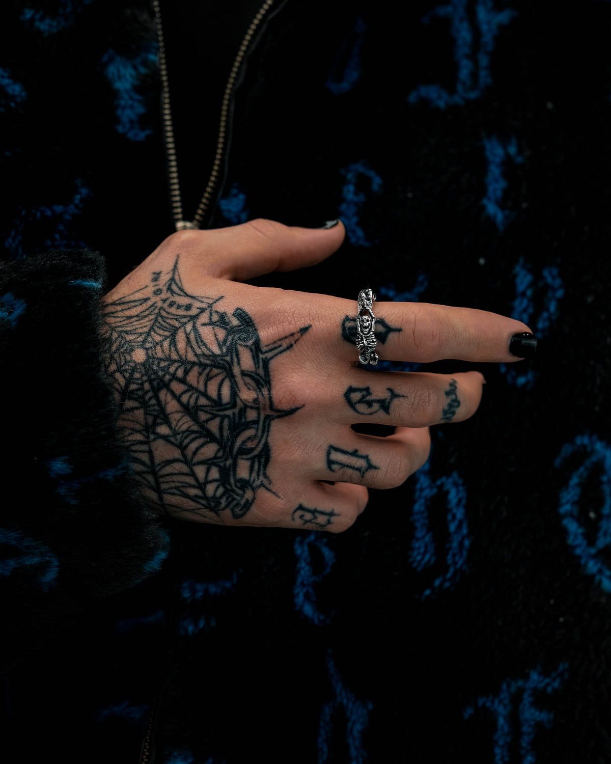 A hand adorned with black nail polish, spider web tattoos, and the Breaking Wheel Ring by Statement Collective is displayed against a dark, blue-patterned jacket. The fingers also feature letter tattoos.