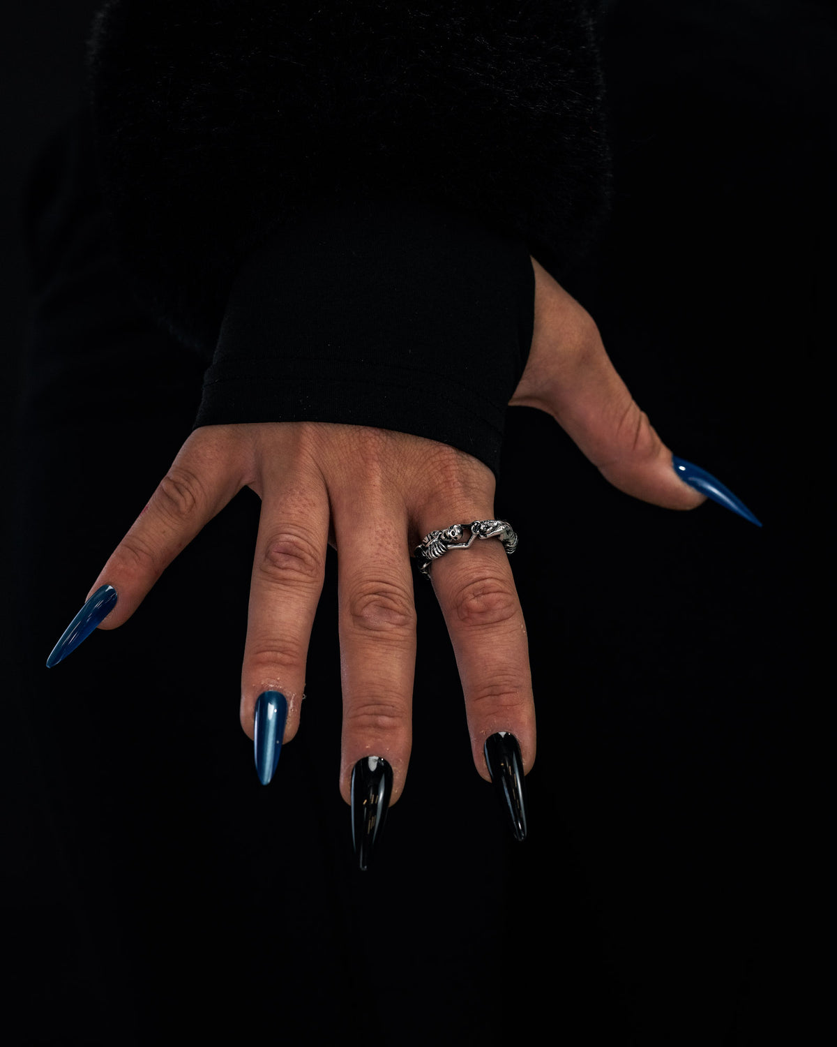 A hand adorned with long, glossy black and blue coffin-shaped nails showcases the Statement Collective Breaking Wheel Ring against a dark background.