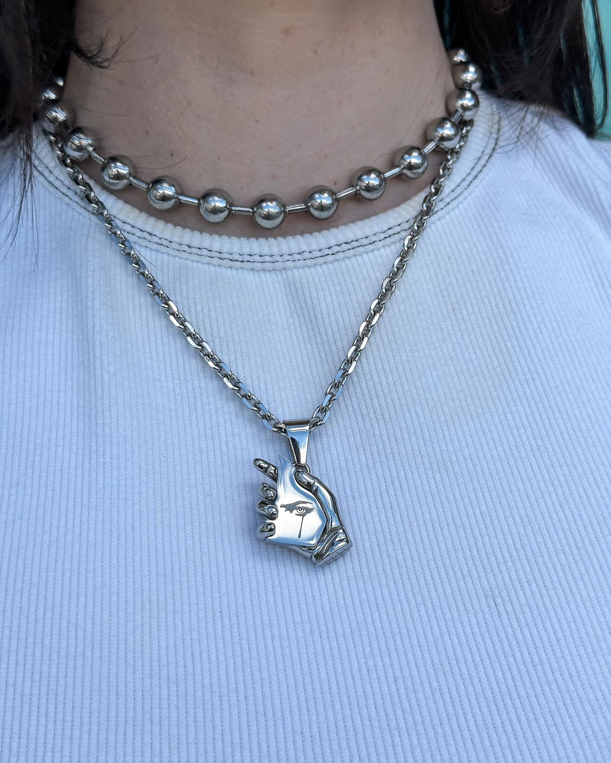 A person wears a ribbed white shirt and two silver necklaces: one with round beads, the other featuring Statement Collective's "The Broken Mirror" Pendant—a hand-shaped piece flashing a peace sign for bold, avant-garde style.