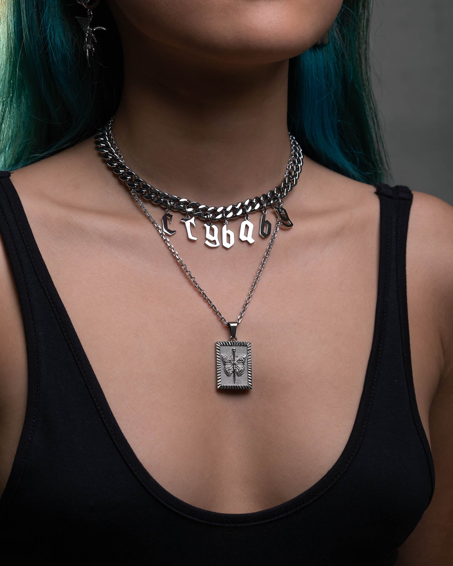 A person with teal hair wears a black tank top and layered silver necklaces—one a chunky chain with letter charms, the other featuring the Butterfly Tag Pendant with an embossed design.