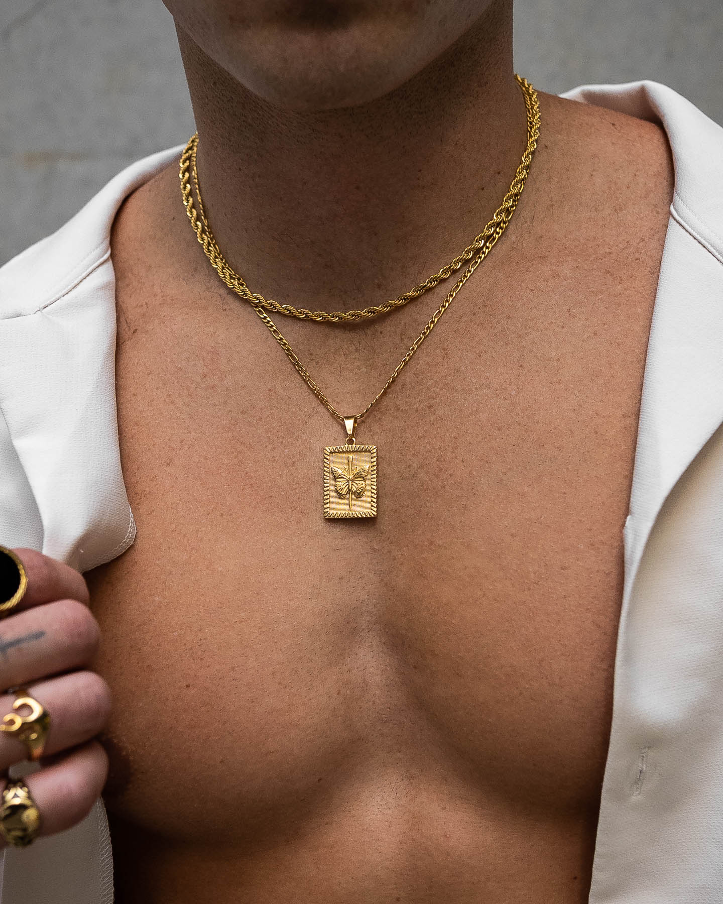 A shirtless person wears two gold chains—one is the Butterfly Tag Pendant (Gold) necklace. Gold rings adorn their fingers, and an open white shirt is draped over their shoulders.