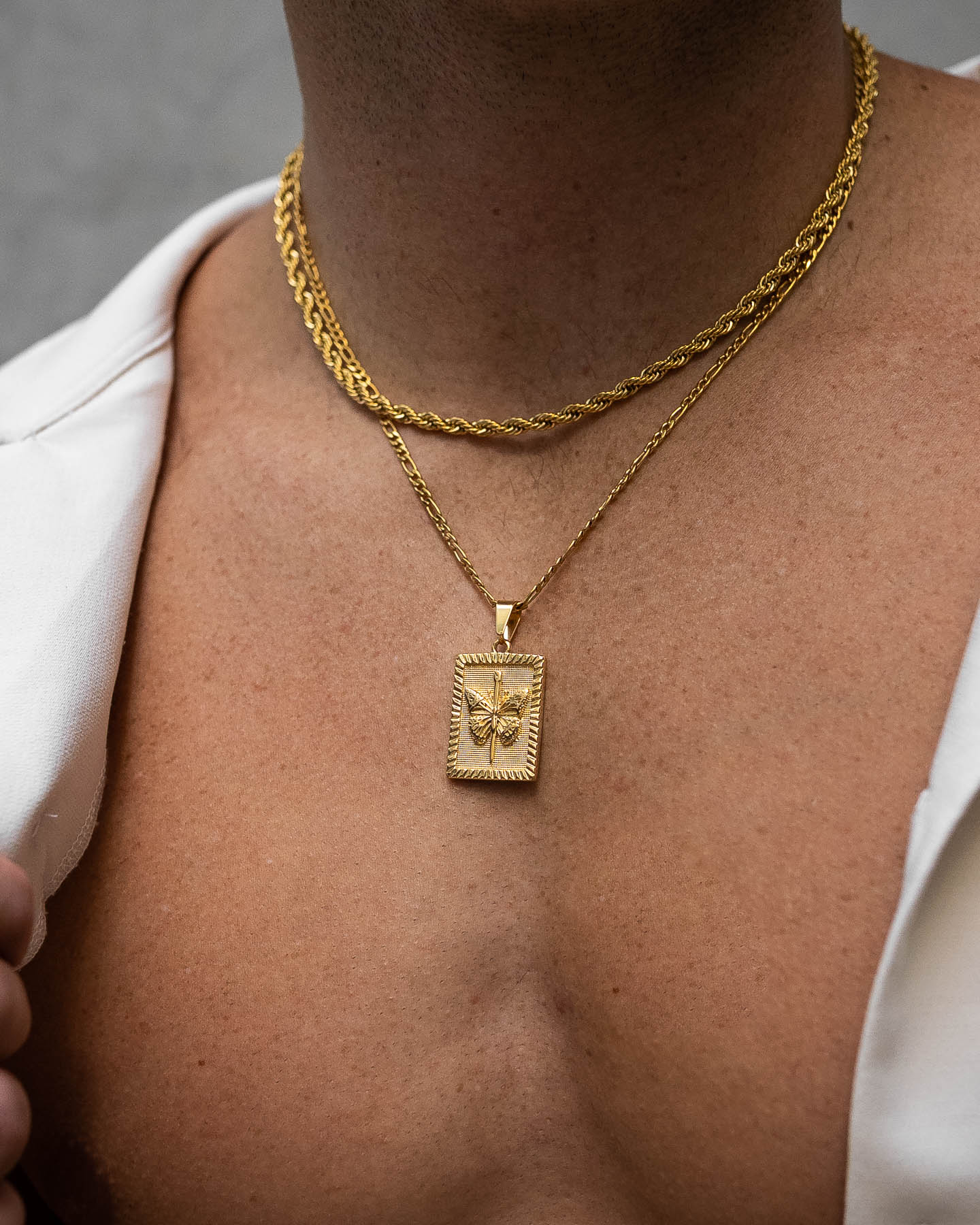 Close-up of a person wearing two gold chains: an 18K gold plated necklace and the Butterfly Tag Pendant (Gold), a rectangular pendant with a raised butterfly design, shown over an open white shirt.