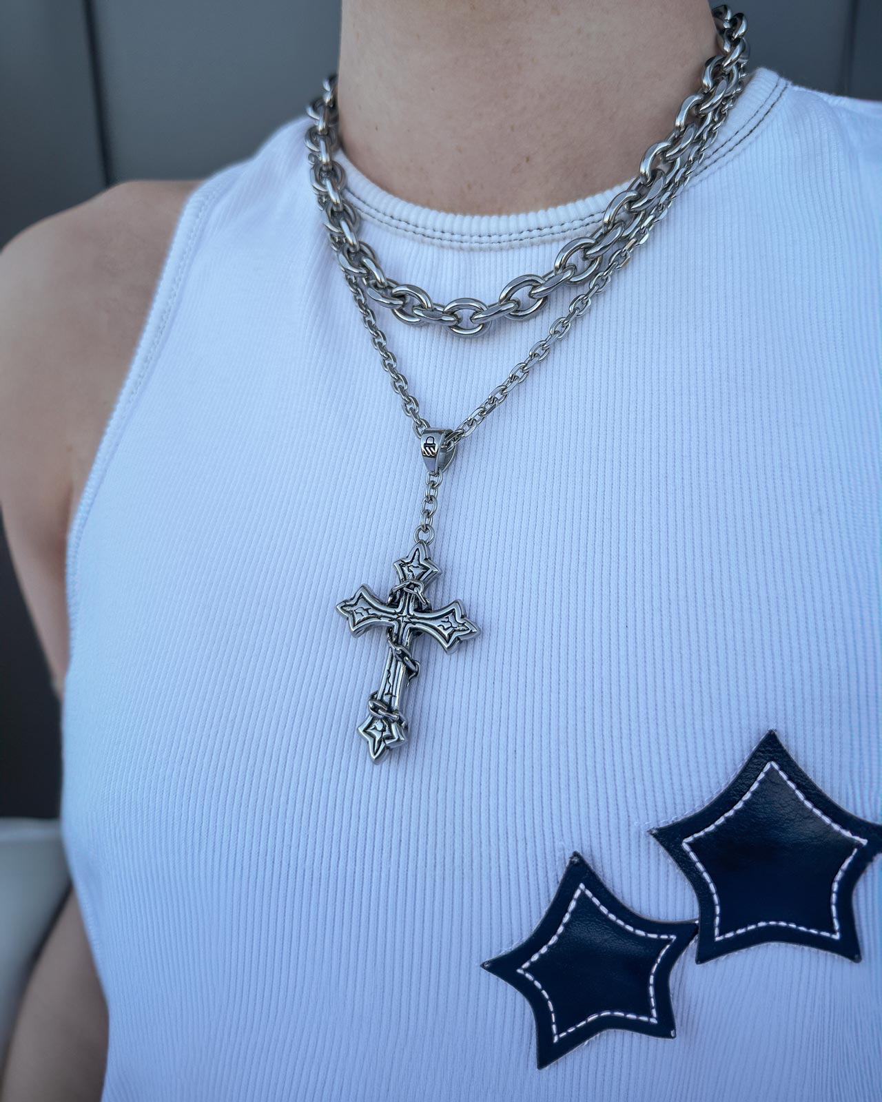 Someone wears a white sleeveless ribbed top with two black star patches and two silver chain necklaces from Statement Collective’s Cable Set (Custom Pendant Choice), featuring a large ornate cross pendant on a Beveled Cable Chain.