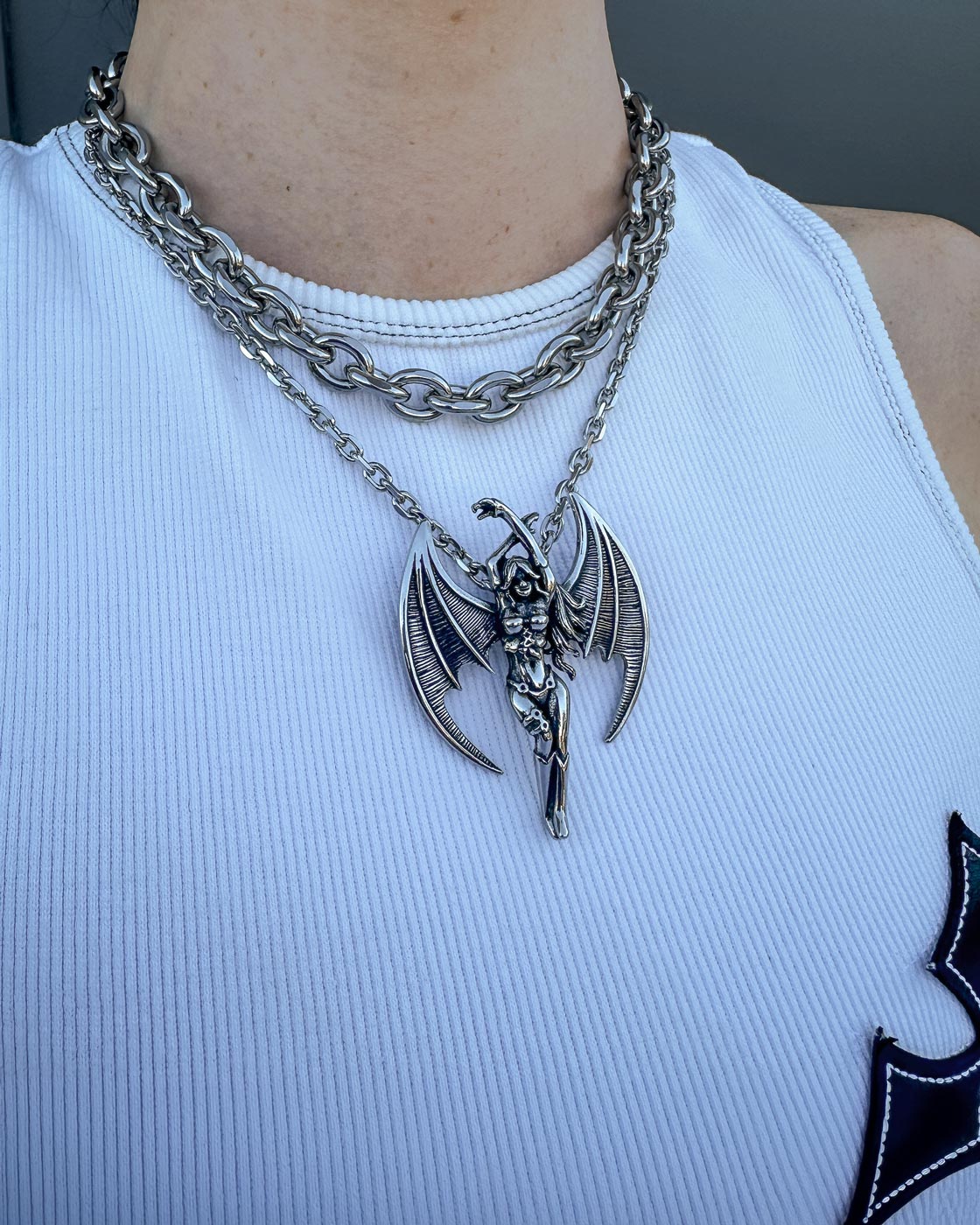 A person wears a white ribbed tank top and Statement Collective's Cable Set—layered stainless steel chain necklaces, one with a large custom winged demon pendant.