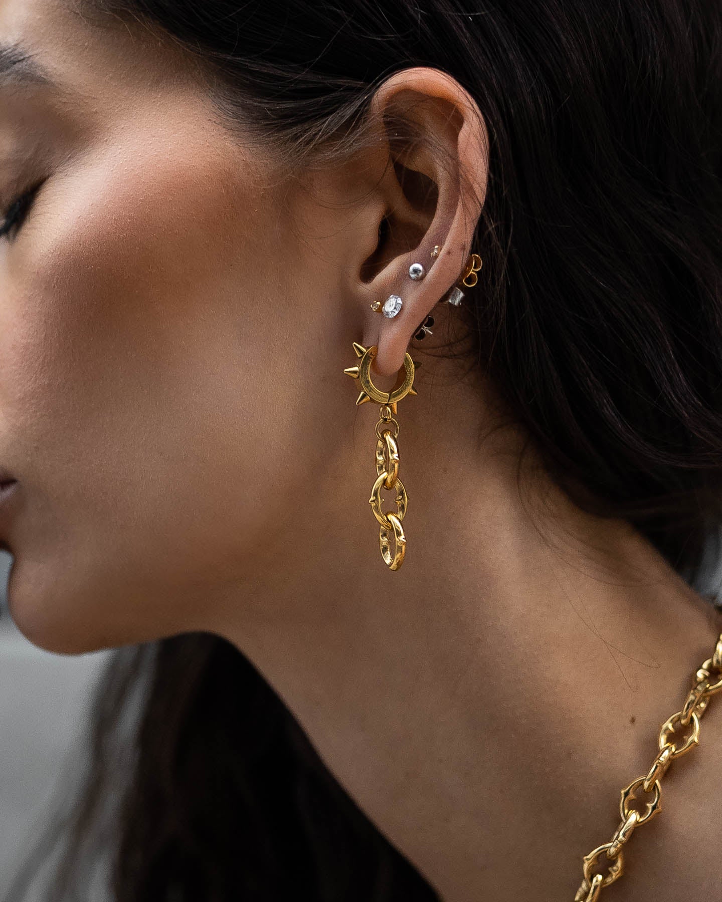 Close-up of a woman’s ear adorned with multiple earrings, featuring "The Cathedral" 9mm Chain Earrings (Gold), along with gold hoops and studs. She also wears a chunky gold chain necklace, and her dark hair is partially visible.