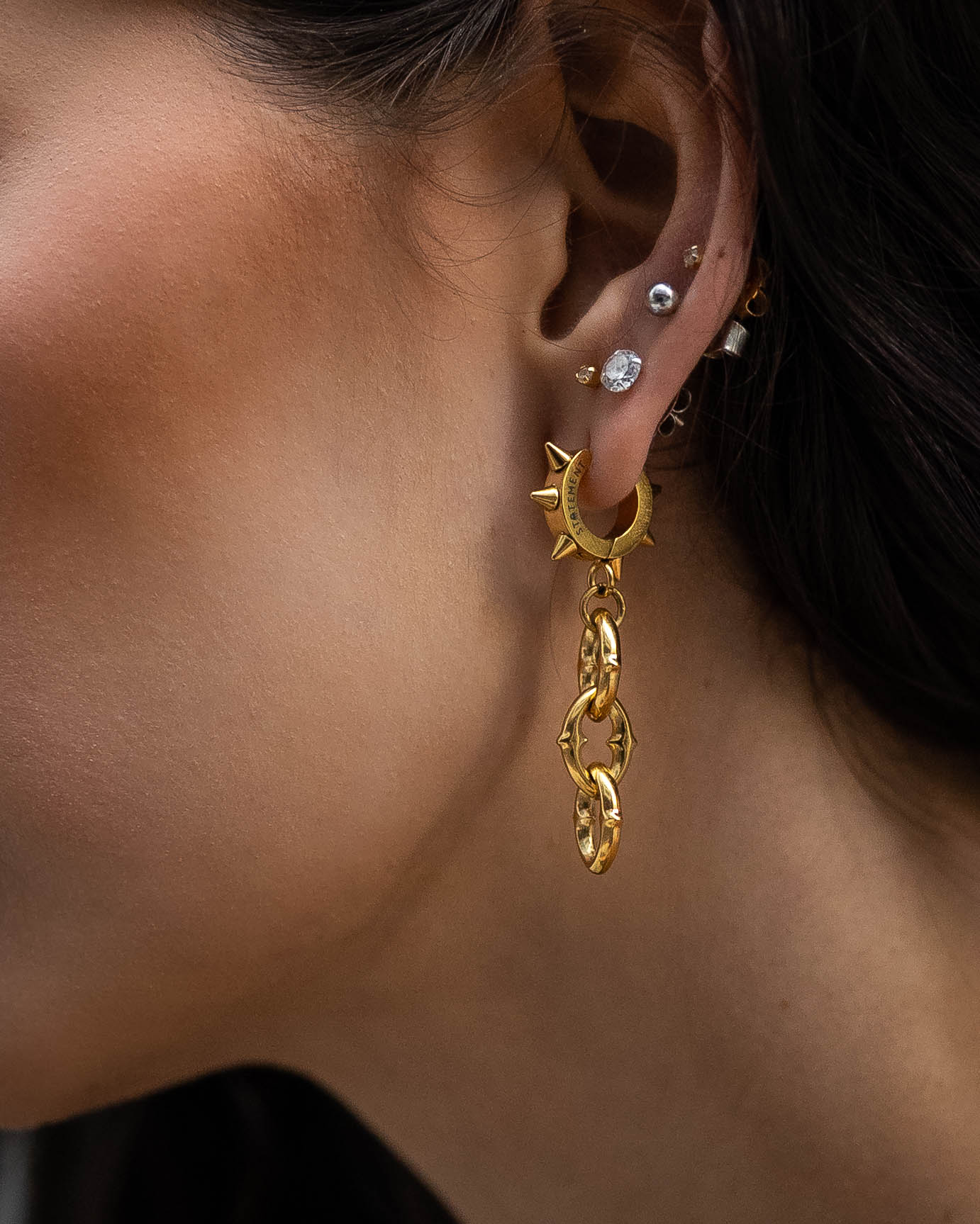 Close-up of a person’s ear with multiple piercings, featuring "The Cathedral" 9mm Chain Earrings in gold, alongside spiked chain earrings and several smaller silver studs and hoops.