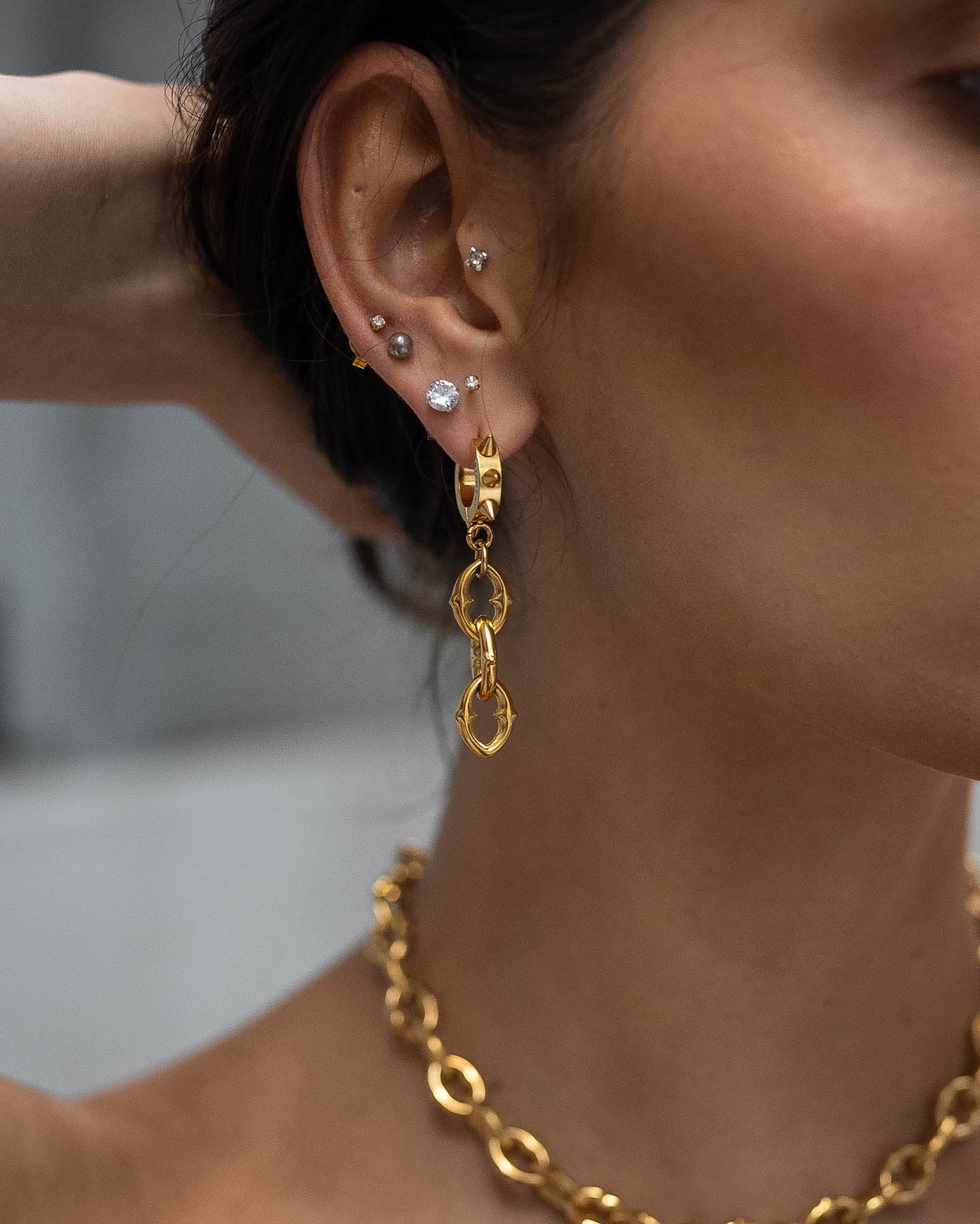 A close-up of a woman’s ear wearing multiple small studs and bold "The Cathedral" 9mm Chain Earrings (Gold), paired with a chunky gold chain necklace. Her skin is smooth, and her dark hair is pulled back.