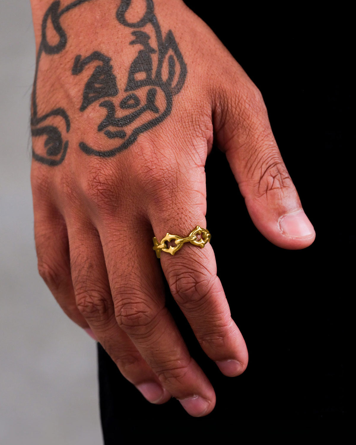 A hand with a cartoon-style devil tattoo wears Statement Collective’s "The Cathedral" Spiked Ring (Gold)—a chunky, twisted 18k gold piece—on the ring finger. The plain dark background highlights its bold elegance.
