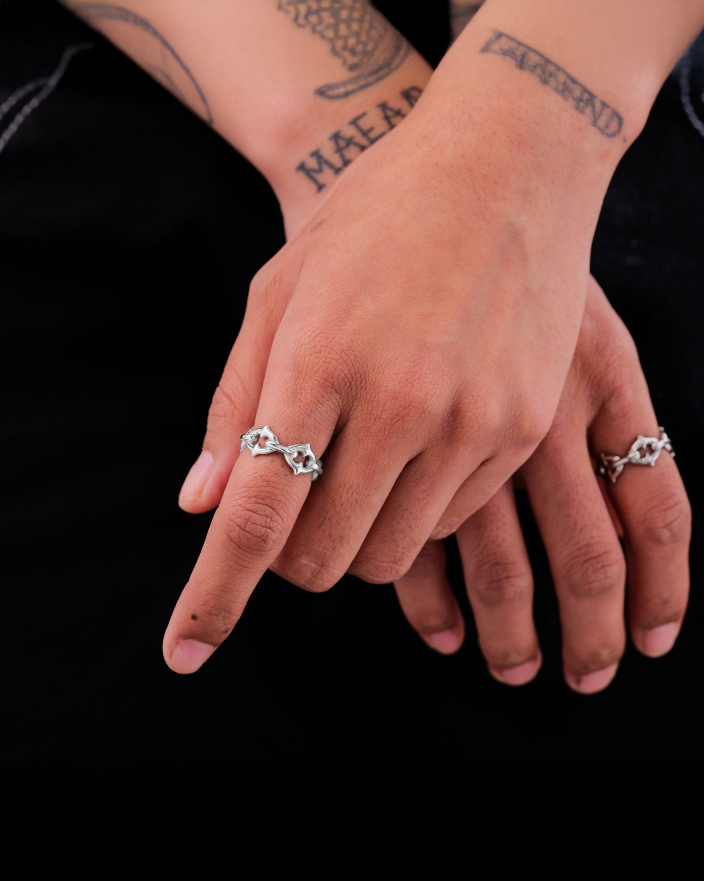 A close-up of two hands with tattooed wrists wearing silver rings, including "The Cathedral" Spiked Ring, with a gothic chain design. The dark background highlights the hands and striking jewelry.