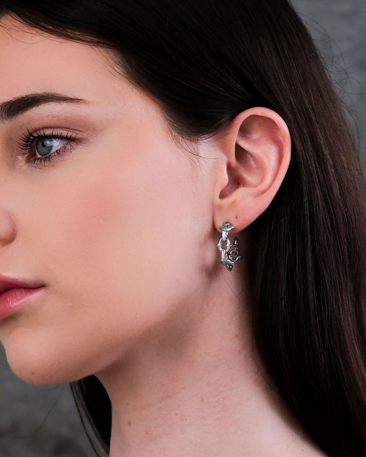 A close-up of a woman’s ear showcases "The Cathedral" Hoop Earrings. She has fair skin, straight dark brown hair, and is set against a neutral background.