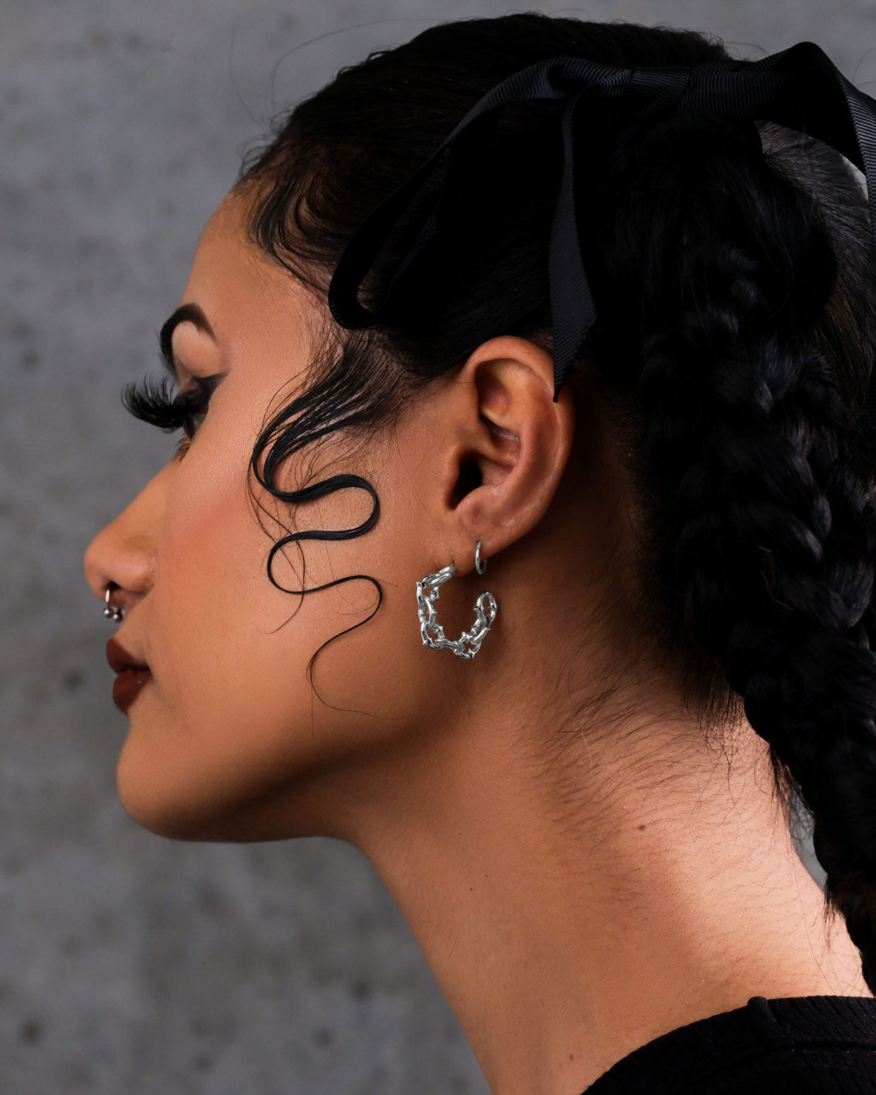 A person with braided hair, a black ribbon, and a nose ring wears "The Cathedral" Hoop Earrings and gothic-inspired accessories. A defined curl graces their cheek against a gray, textured background.
