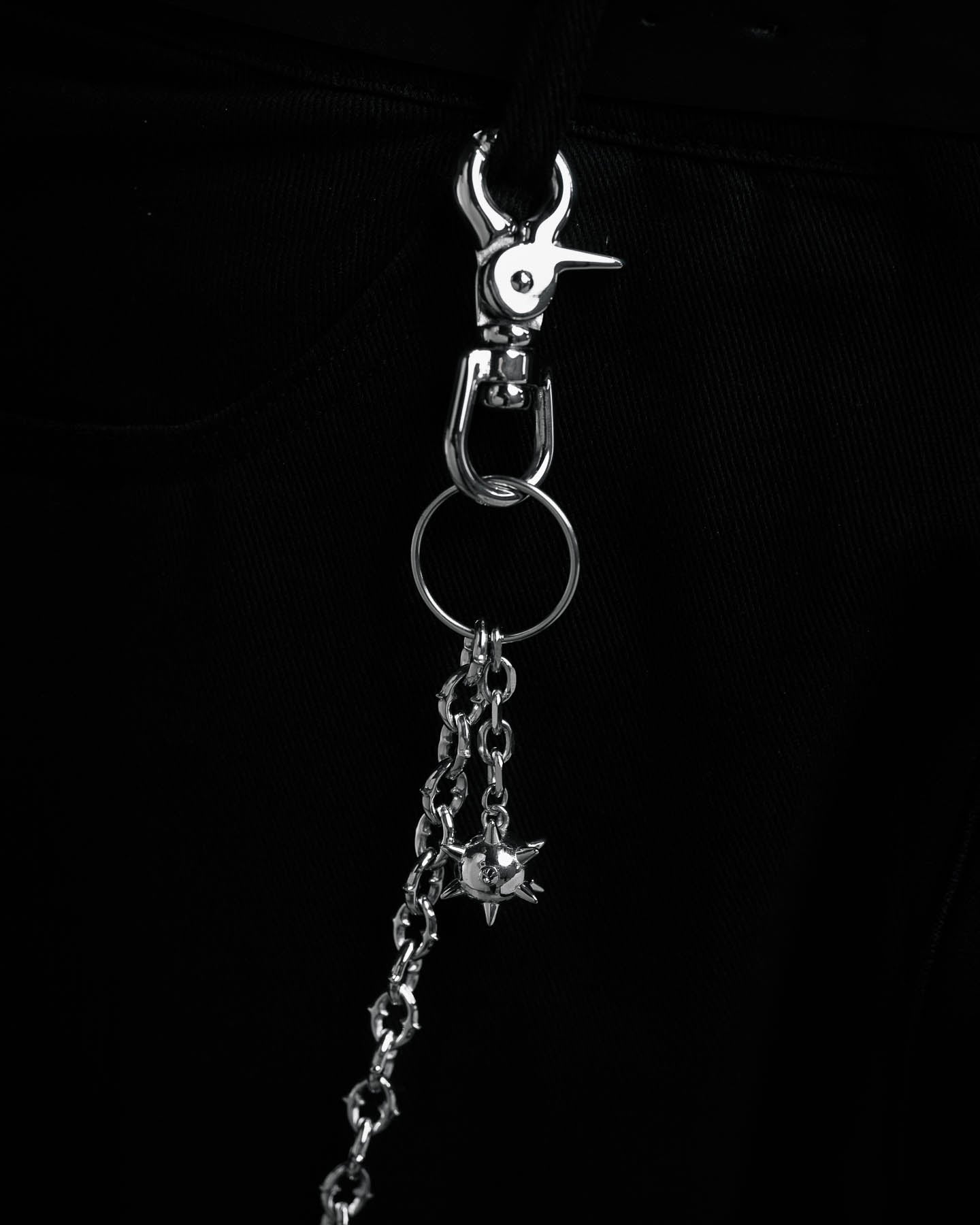 A close-up of "The Cathedral" 9mm Spiked Wallet Chain with a ball charm, metal clip, and key ring, hanging from black fabric—possibly pants. The dark, monochromatic look adds to its edgy streetwear style.