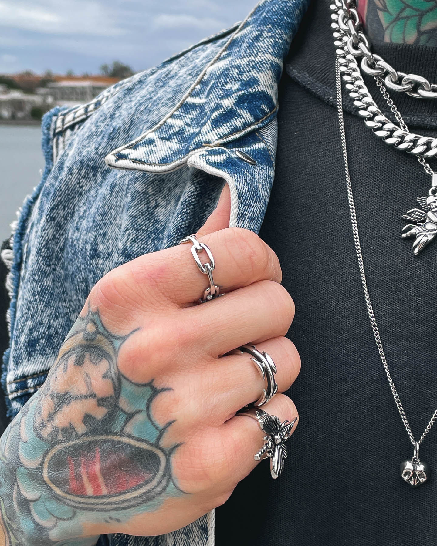 A tattooed hand wearing the Chained Ring holds the collar of a blue denim jacket. Silver necklaces and a black shirt complete the look, with water and city buildings softly blurred in the background.