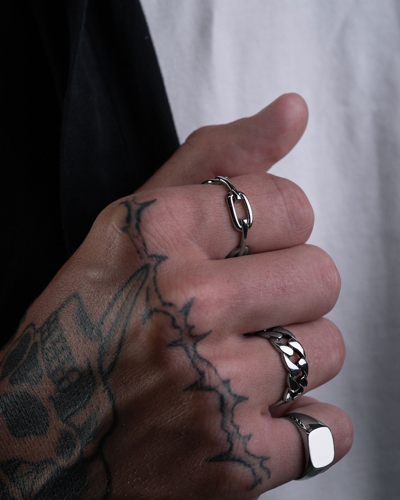 A close-up of a hand with black ink tattoos wearing the Chained Ring and two band rings, set against a background showing part of a black sleeve and white shirt.