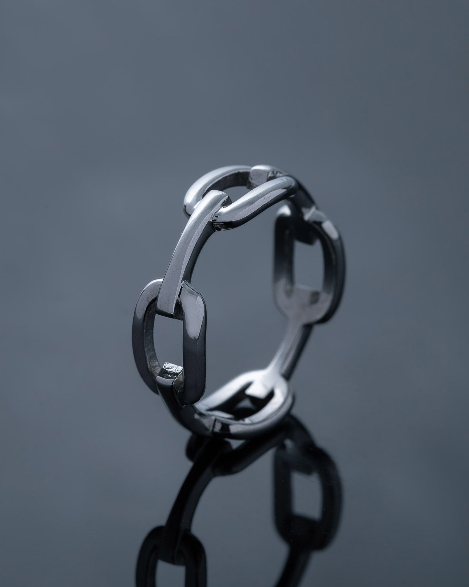The Chained Ring, crafted in silver with interlocking links, stands upright on a glossy dark surface that mirrors its bold shape—a striking piece ideal for stacking.