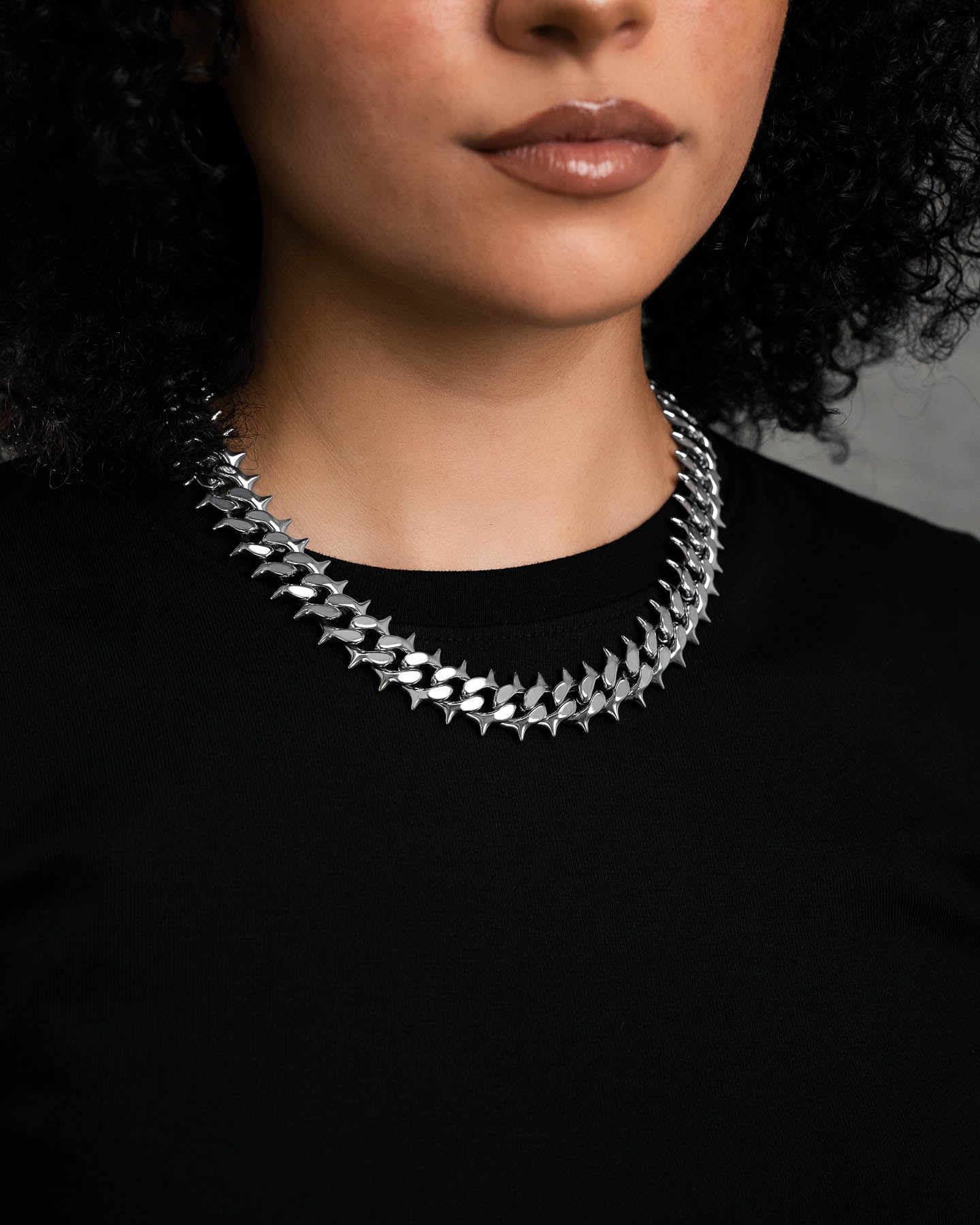 A woman with curly hair wears the "Chainsaw" Spiked Cuban Chain over a black top. Only her lower face, neck, and upper chest are visible against a neutral, blurred background.