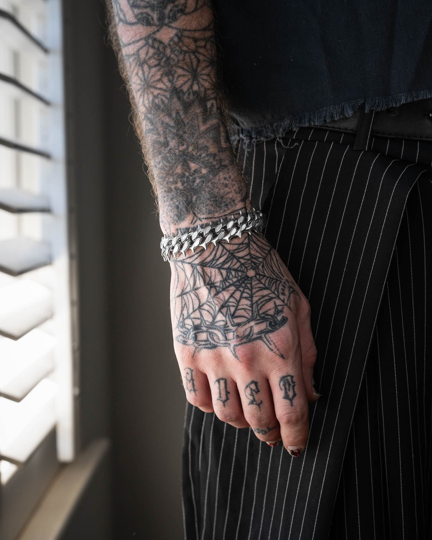 A tattooed hand with a spiderweb design wears the "Chainsaw" Spiked Cuban Bracelet, resting against pinstriped pants by a window with blinds.