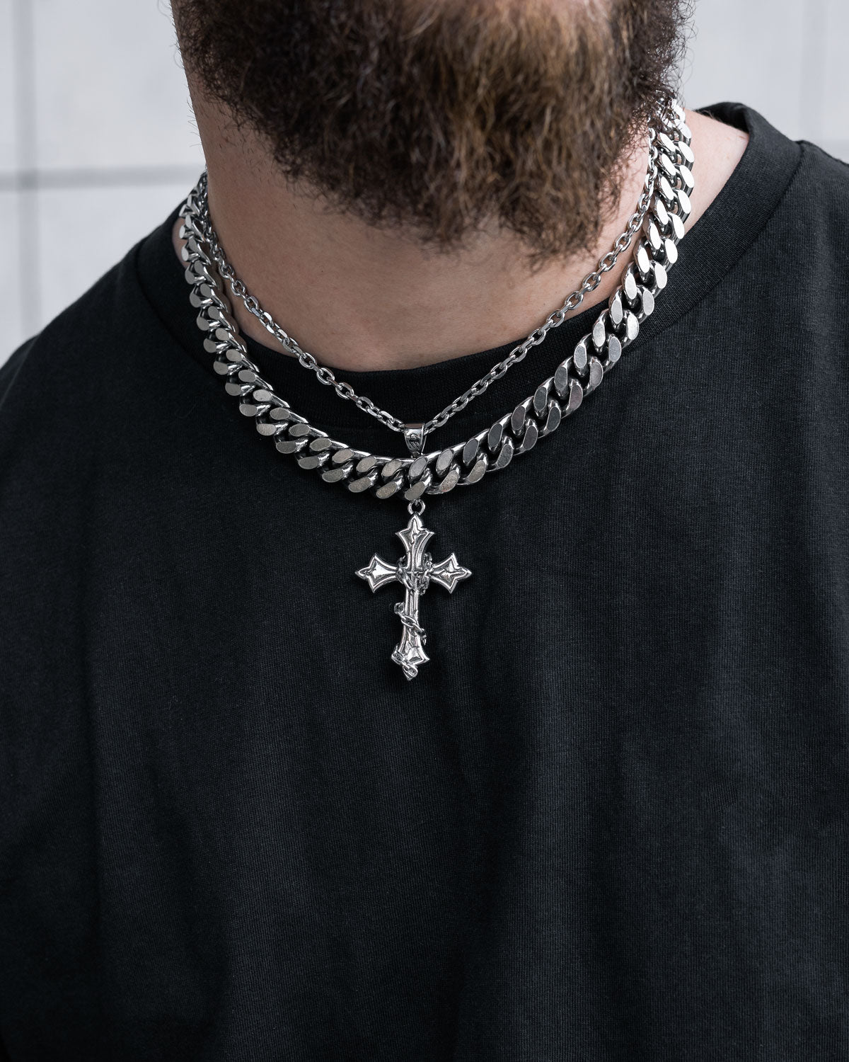 A man with a brown beard in a black shirt wears the Statement Collective Chunky Ruins Set—a layered necklace with two silver chains, including a Cuban link chain and stainless steel cross pendant—against light-colored tile background.