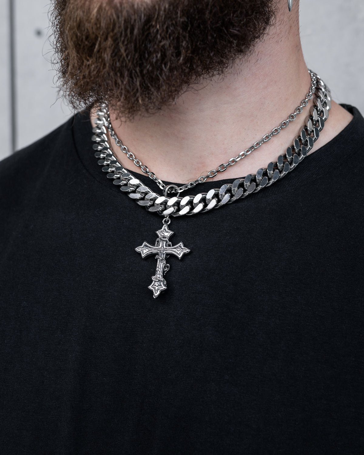 A person with a partial beard wears a black shirt and Statement Collective's Chunky Ruins Set—a layered necklace set featuring silver chains, including a Cuban link and a stainless steel detailed cross pendant.