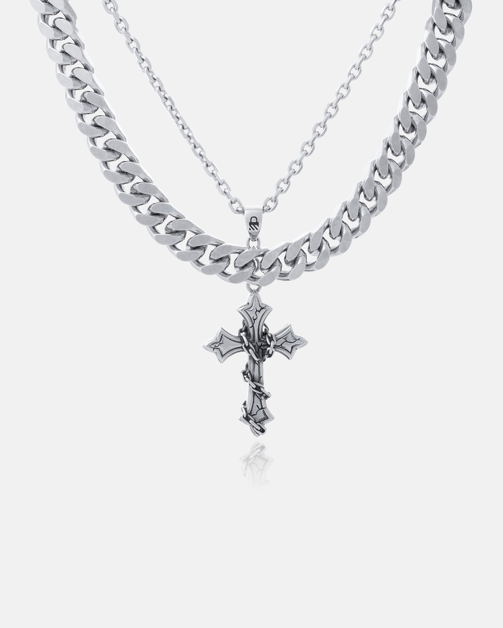 The Chunky Ruins Set features two layered silver chains: a thick Cuban Link and a longer, thinner chain with an ornate Ruins Cross Pendant. Crafted from stainless steel, these striking pieces stand out against a plain white background.