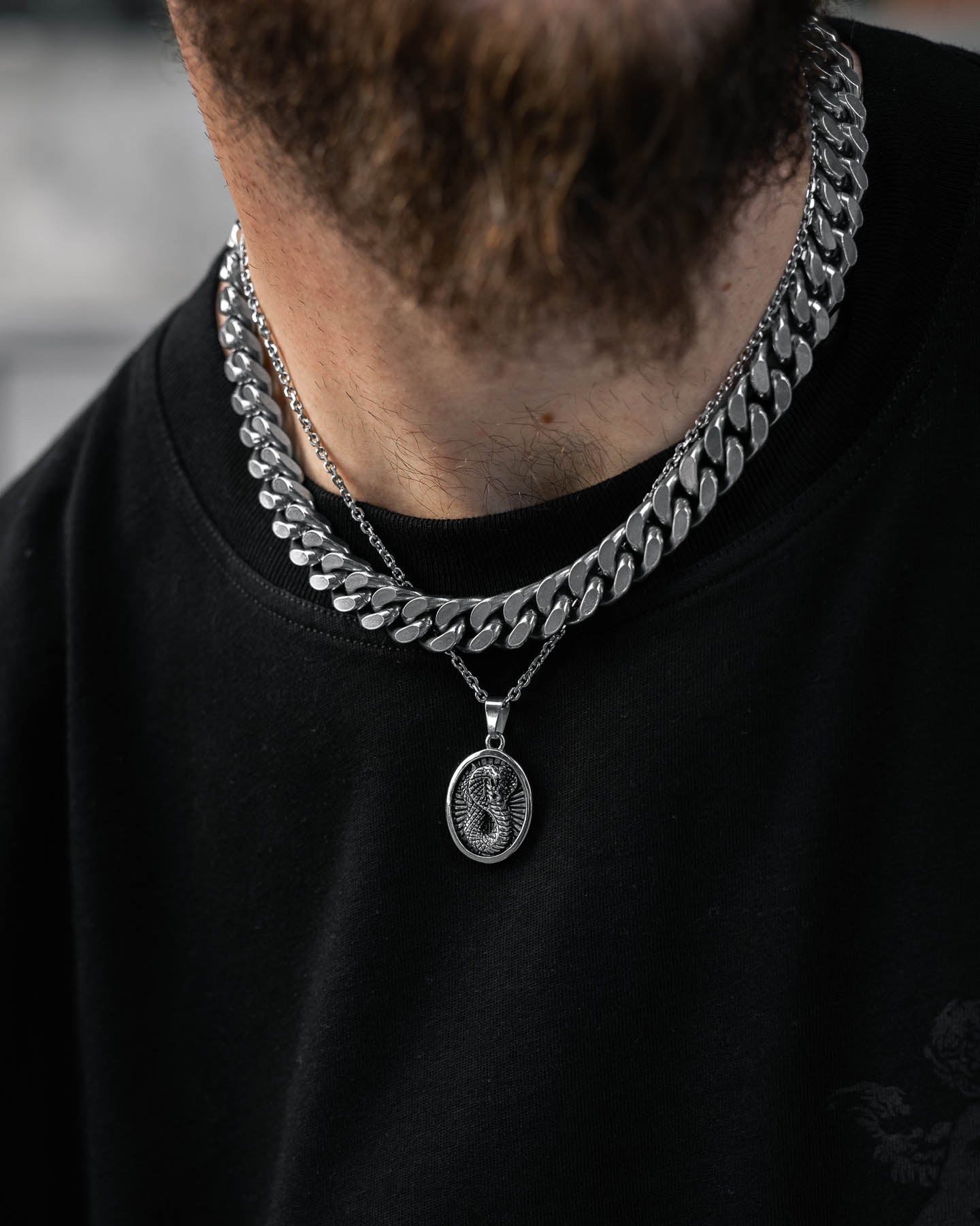 A close-up shows a person wearing two silver chains—a thick, flat one and a thinner chain with the Snake Charmer Pendant, an oval talisman with an engraved design—over a black shirt. The lower face and beard are partially visible.