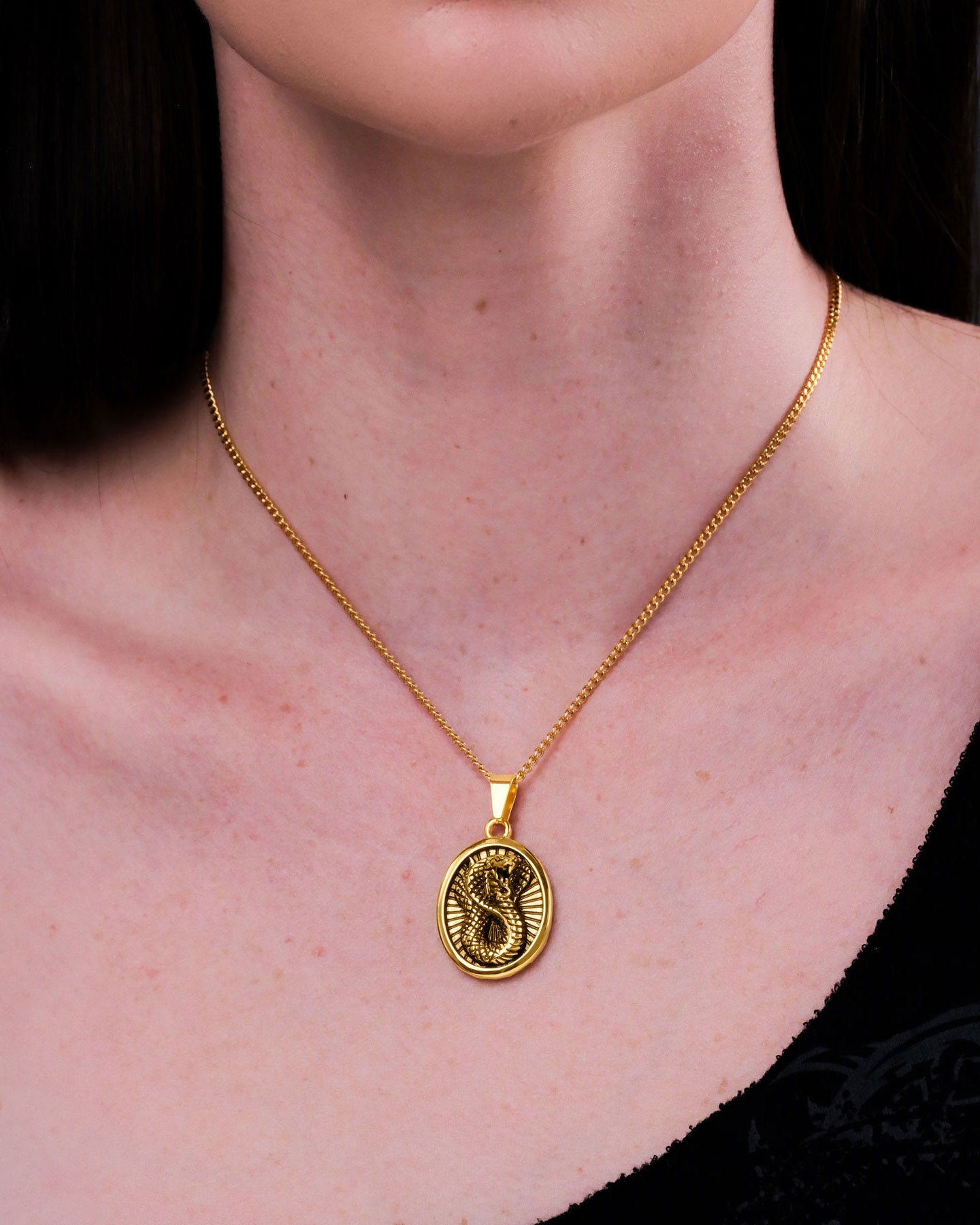 A close-up of a person wearing the Snake Charmer Pendant (Gold), a round 18k gold necklace with a raised snake design, visible above their black top, highlighting their skin and collarbone.