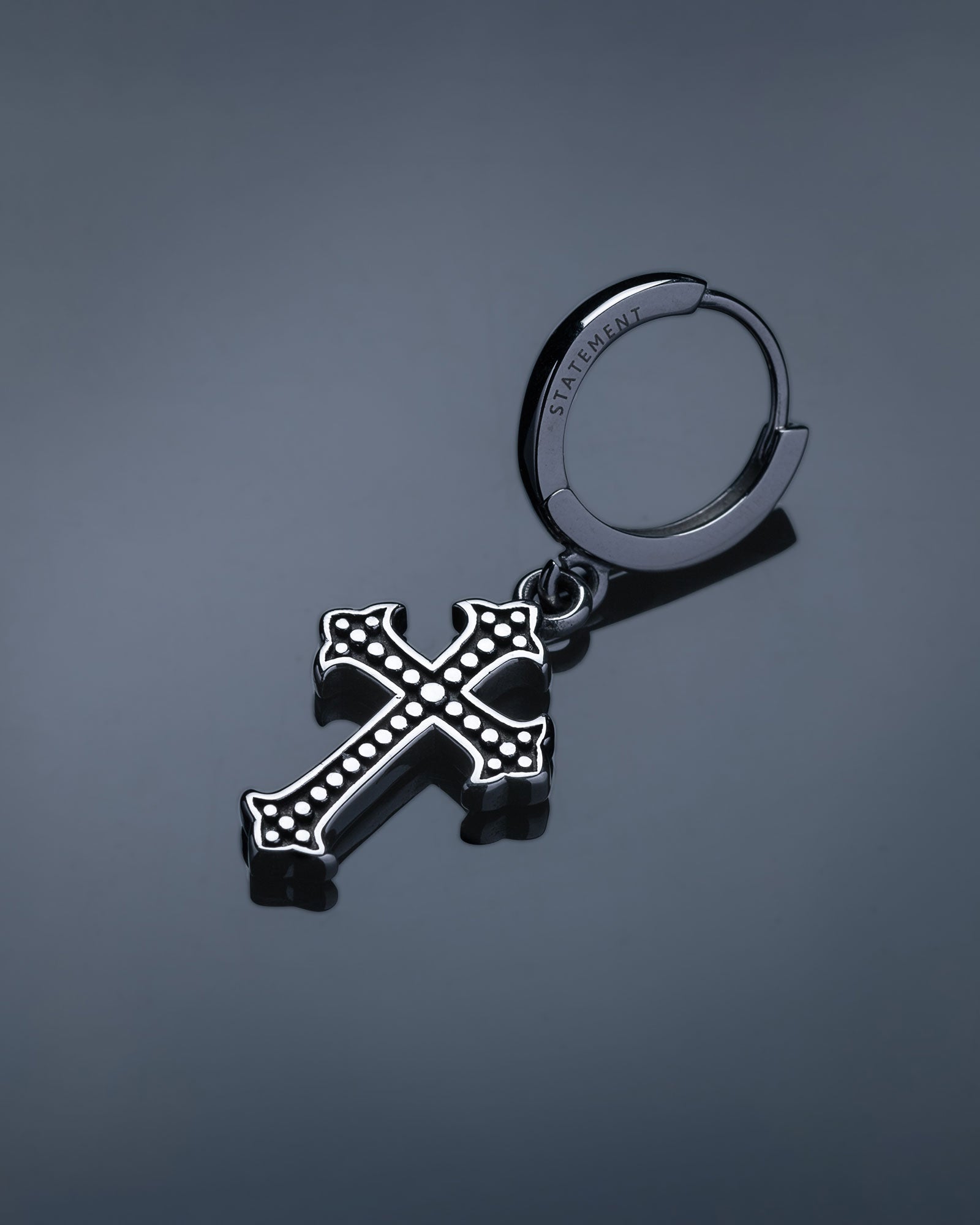 The French Cross Earring is a bold men's silver hoop with a black and silver cross pendant featuring raised dots and "STATEMENT" engraved on the hoop, shown on a smooth gray background.