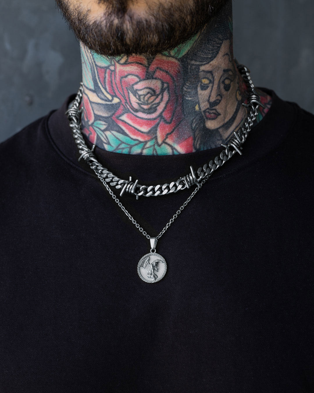 A person with colorful neck tattoos—a rose and a woman's face—wears a black shirt and the Statement Collective Crucifix Set: a chunky barbed wire necklace paired with another featuring a round fallen angel medallion.