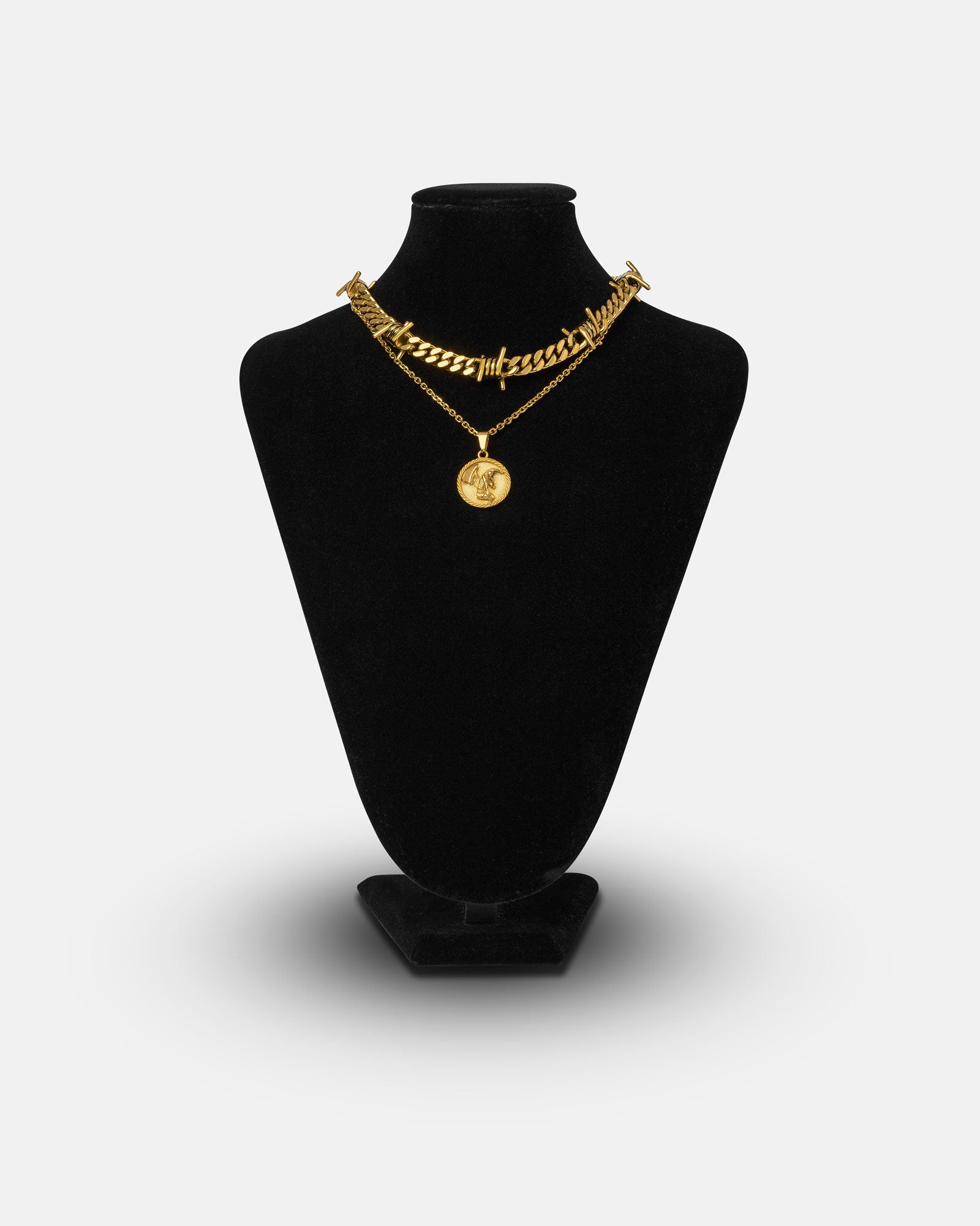 A black velvet necklace stand displays two gold necklaces from the Crucifix Set (Gold): a chunky barbed wire Cuban chain and a finer 18k gold necklace with a round crucifix pendant, against a plain white background.
