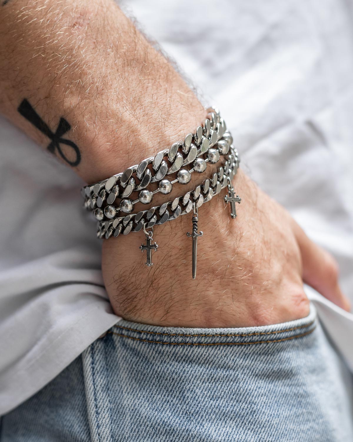 A person wears the Statement Collective "Crusader" 9mm Cuban Bracelet with cross and sword charms, paired with light blue jeans and a white shirt. A black forearm tattoo is visible as their hand rests in their pocket.