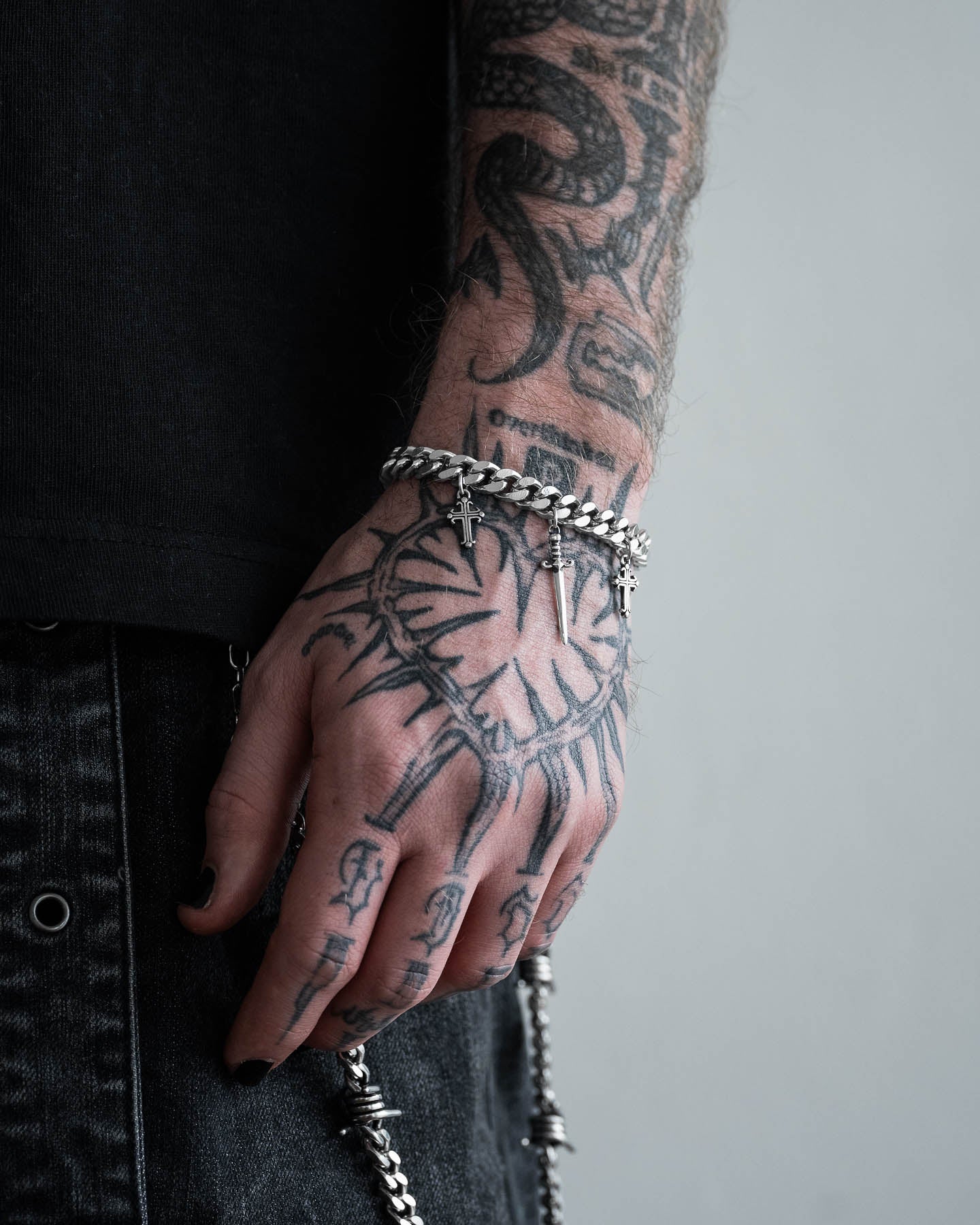 A tattooed hand, covered in intricate designs, rests at the person's side, adorned with the "Crusader" 9mm Cuban Bracelet. Dressed in dark clothing and denim jeans, the bold silver chain completes their look.