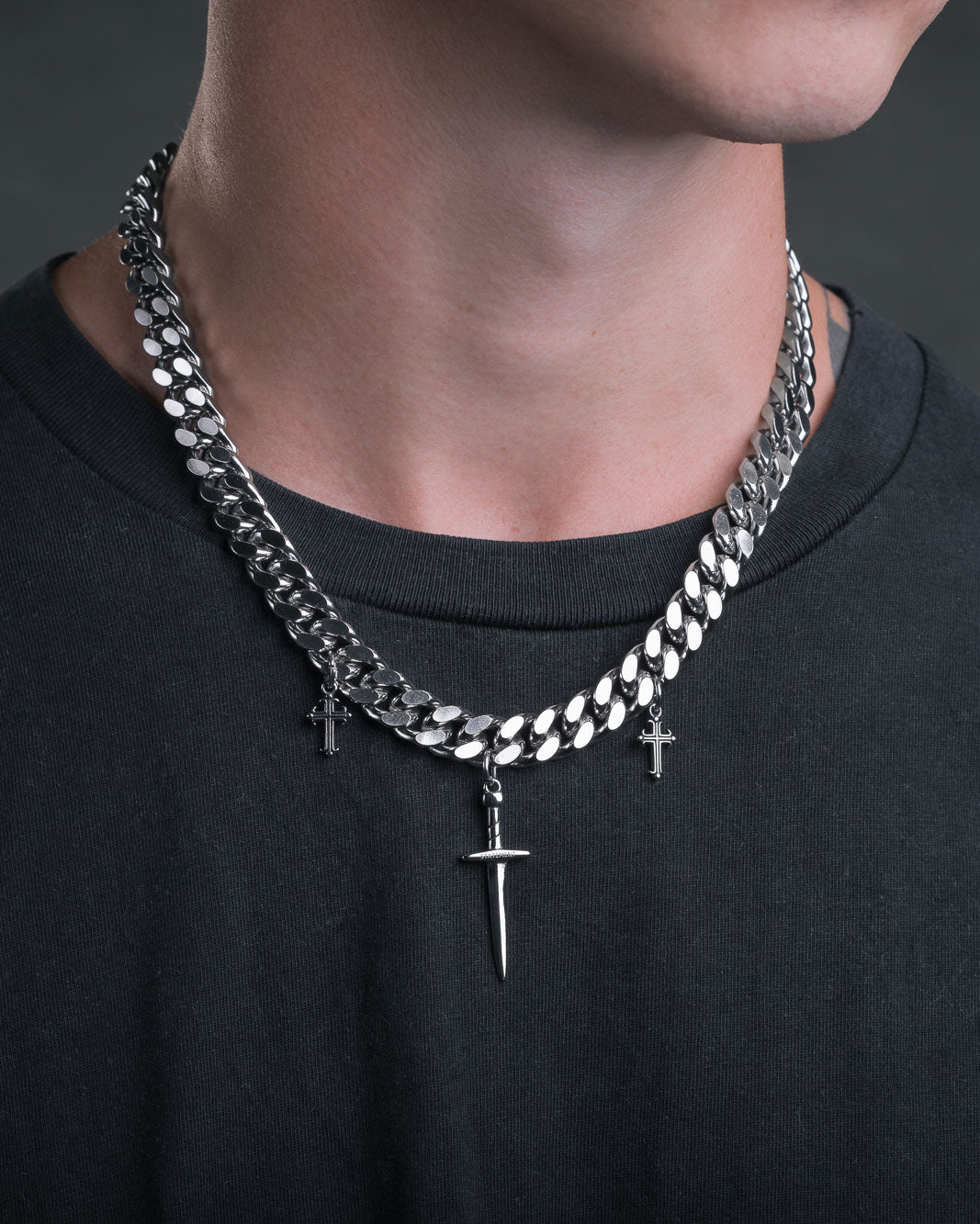 Wearing a black shirt, the person sports two silver chain necklaces: a punk-style one with a large spike pendant and the "Crusader" 11mm Cuban Chain featuring two small cross pendants, all set against a dark background.